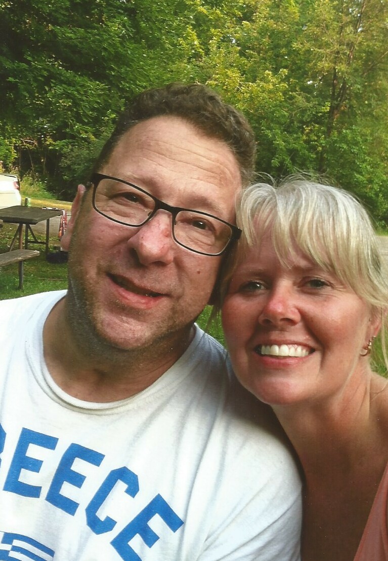 A smiling couple taking a selfie outdoors in a park with green trees in the background.