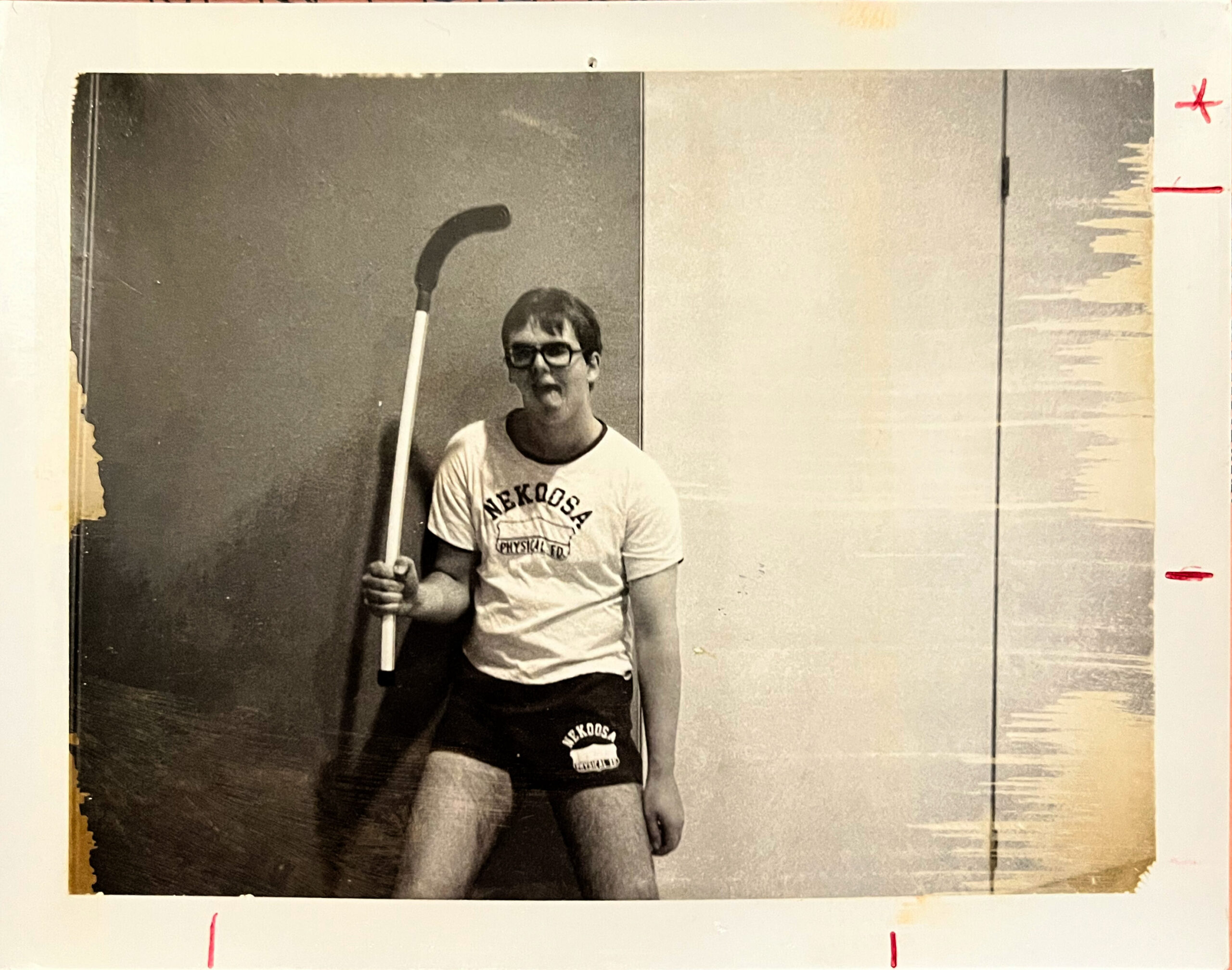 Young man wearing glasses in a gym, holding a curved sports stick over his shoulder, wearing a Nebraska physical education shirt.