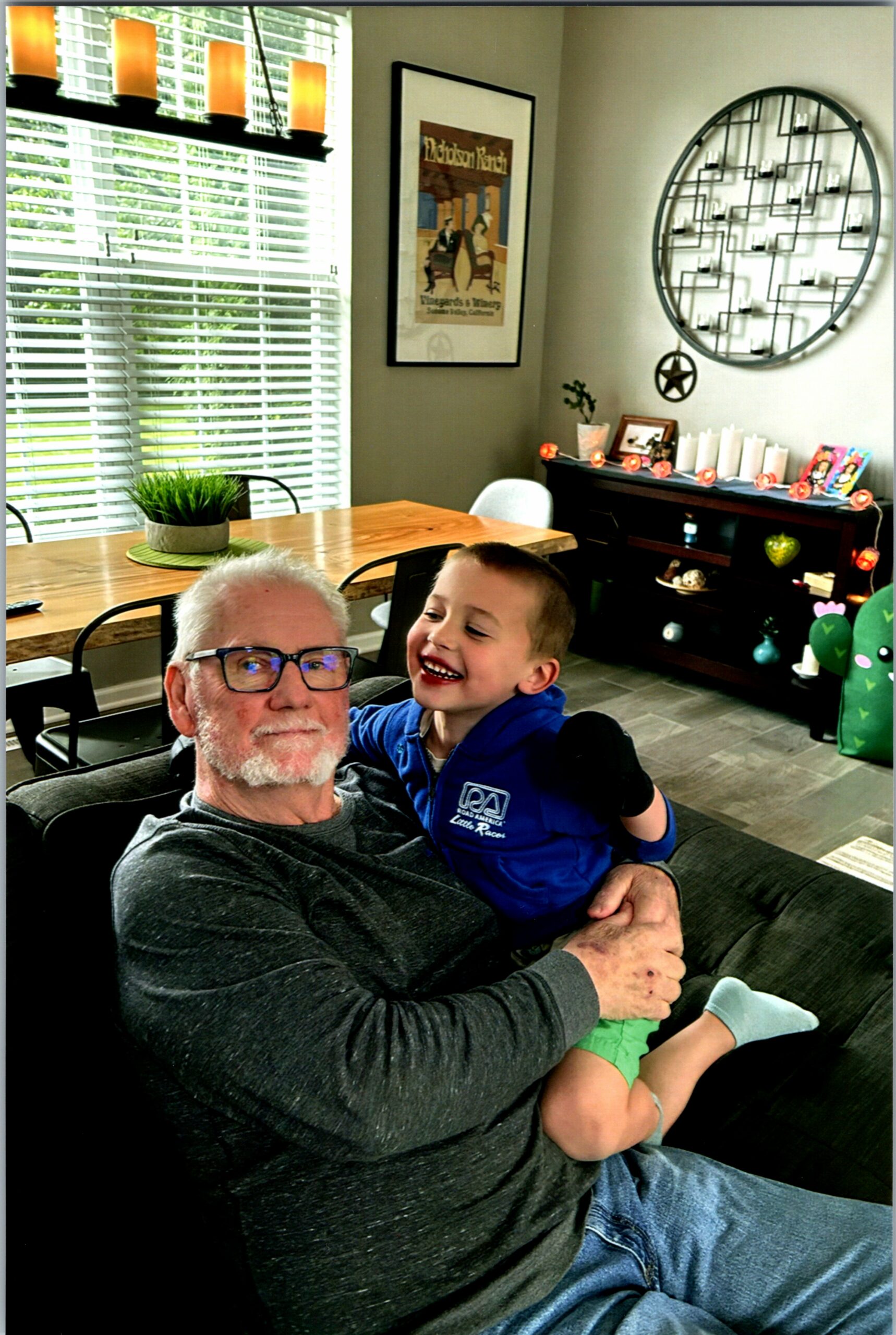 Grandfather and young child smiling together on a sofa in a cozy, sunlit living room with decor in the background.
