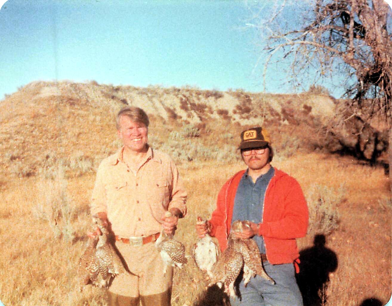 Two hunters stand in a dry field holding several grouse after a hunting trip in bright sunlight.