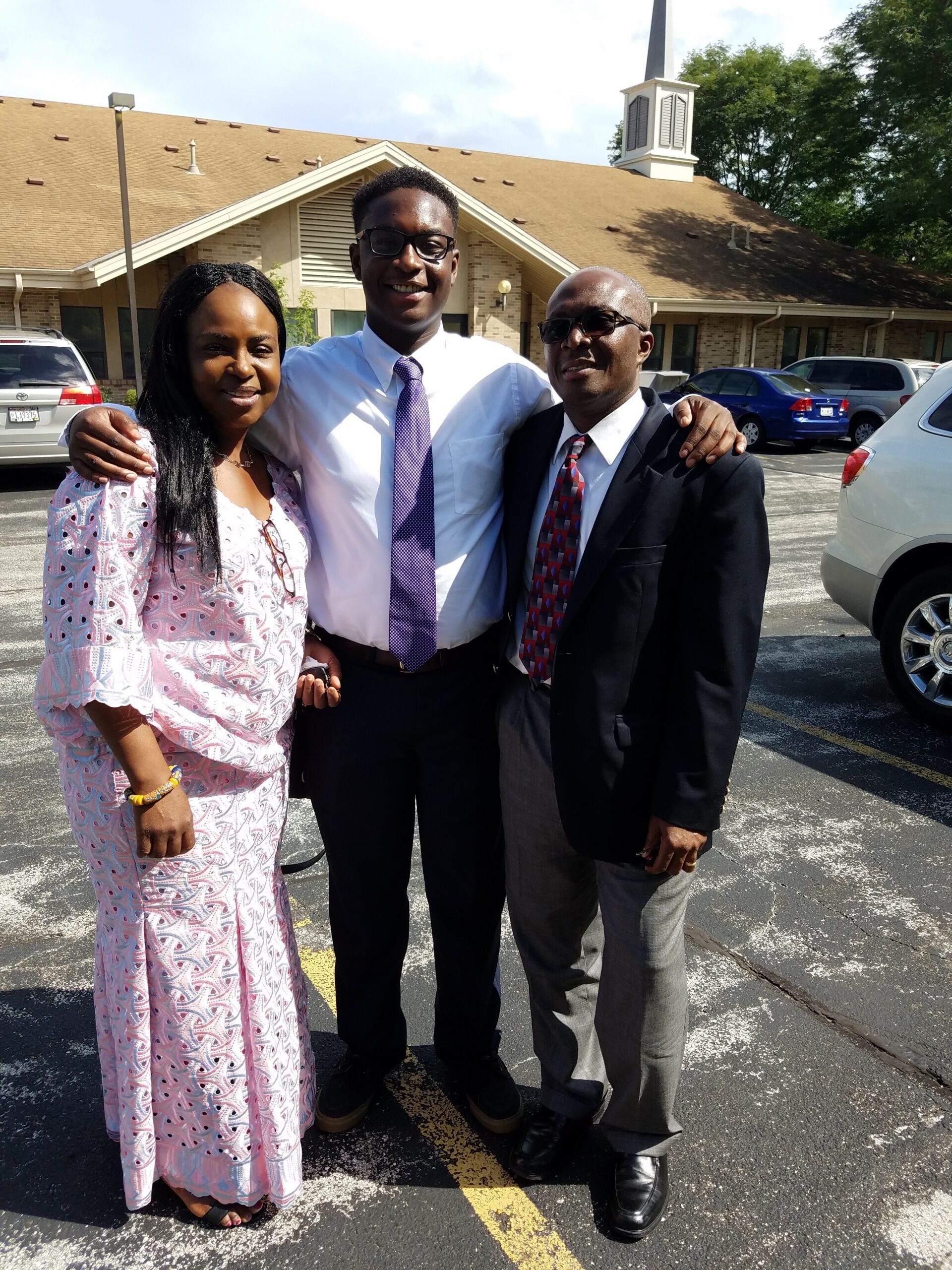 Three adults pose together in a parking lot outside a brick building with a steeple, smiling at the camera; two men in suits and a woman in a pink patterned dress with a white top.