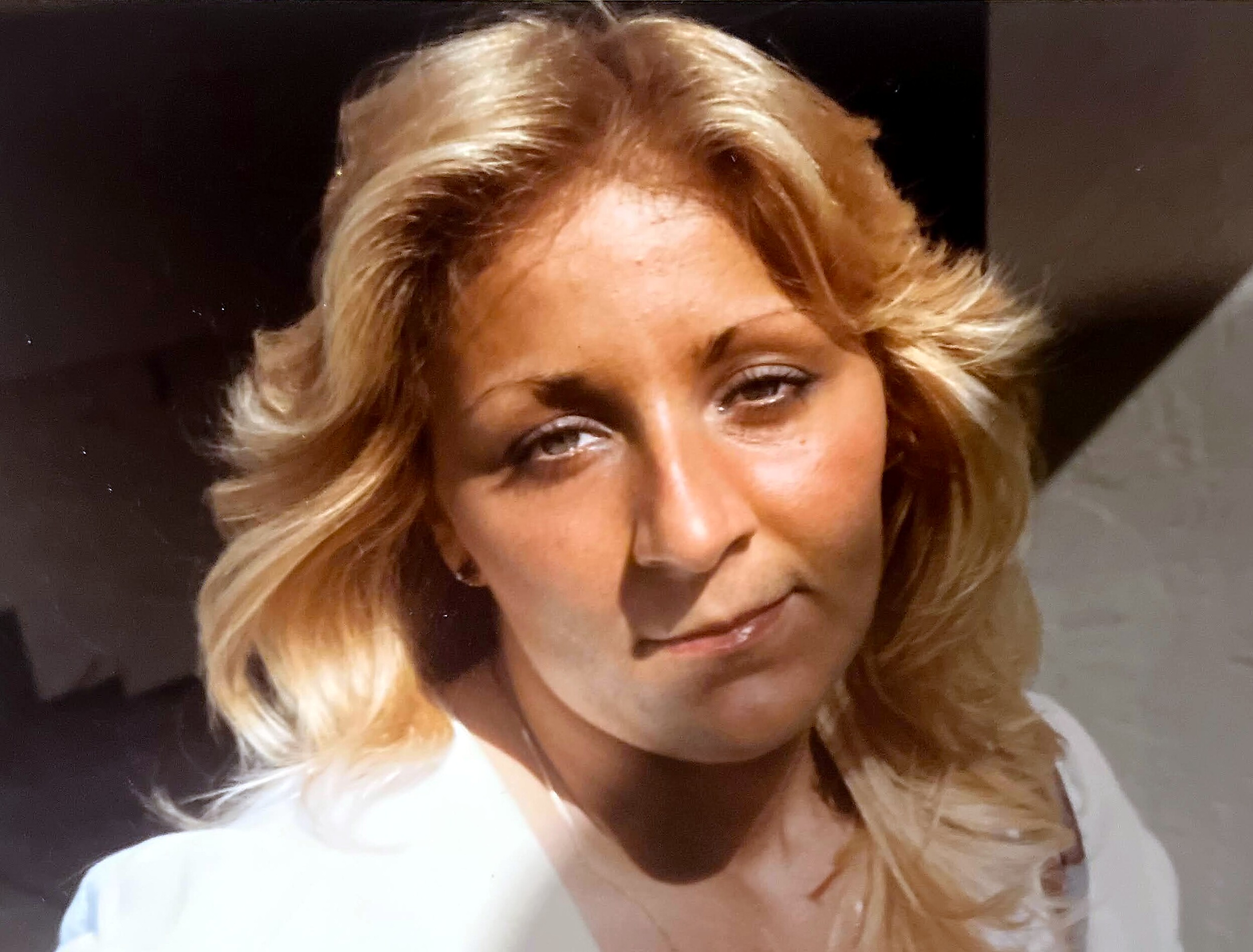 Close-up portrait of a woman with blonde, wavy hair wearing a white blouse, facing the camera.