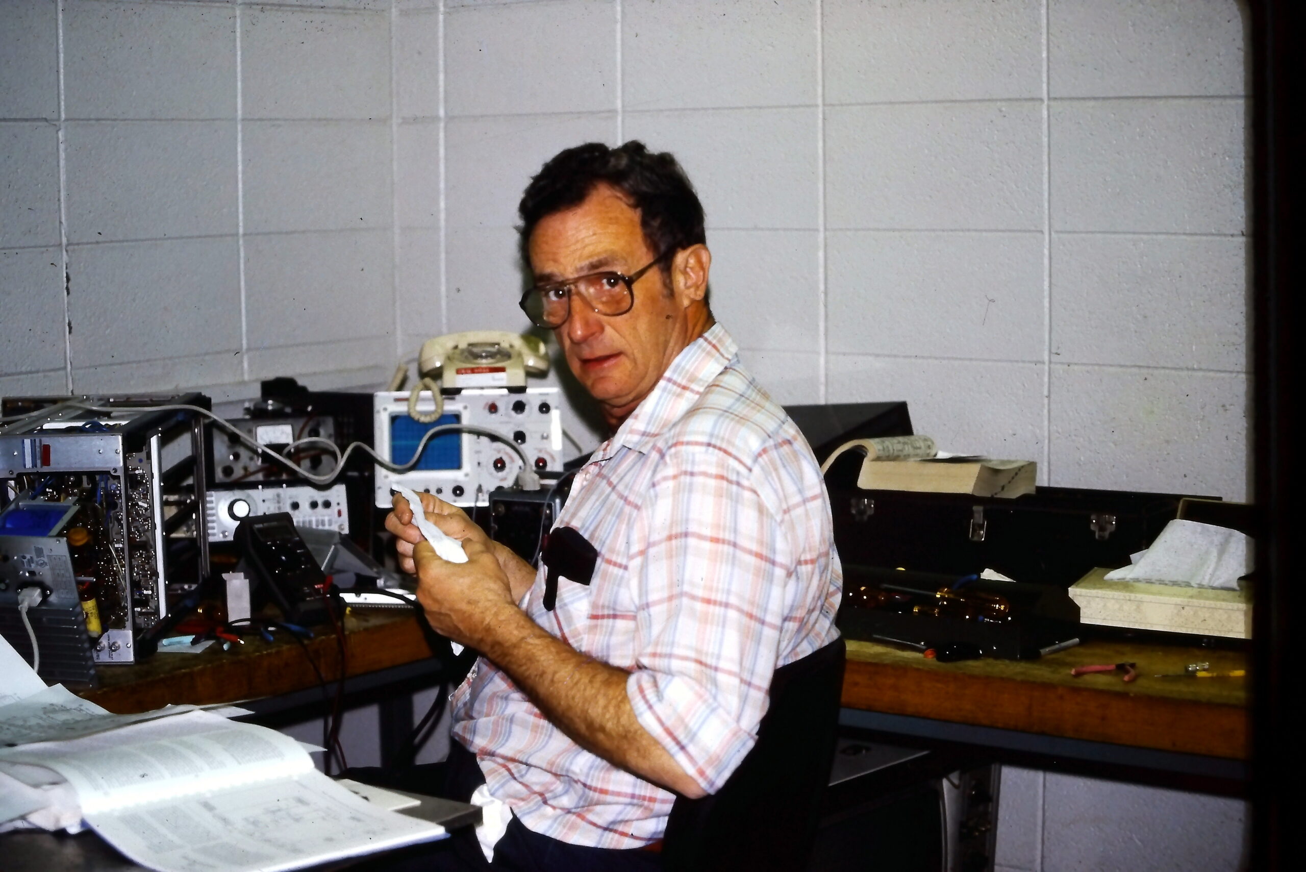 Man with glasses wearing a plaid shirt sits at a cluttered workbench repairing electronic equipment, holding a tissue or cloth.