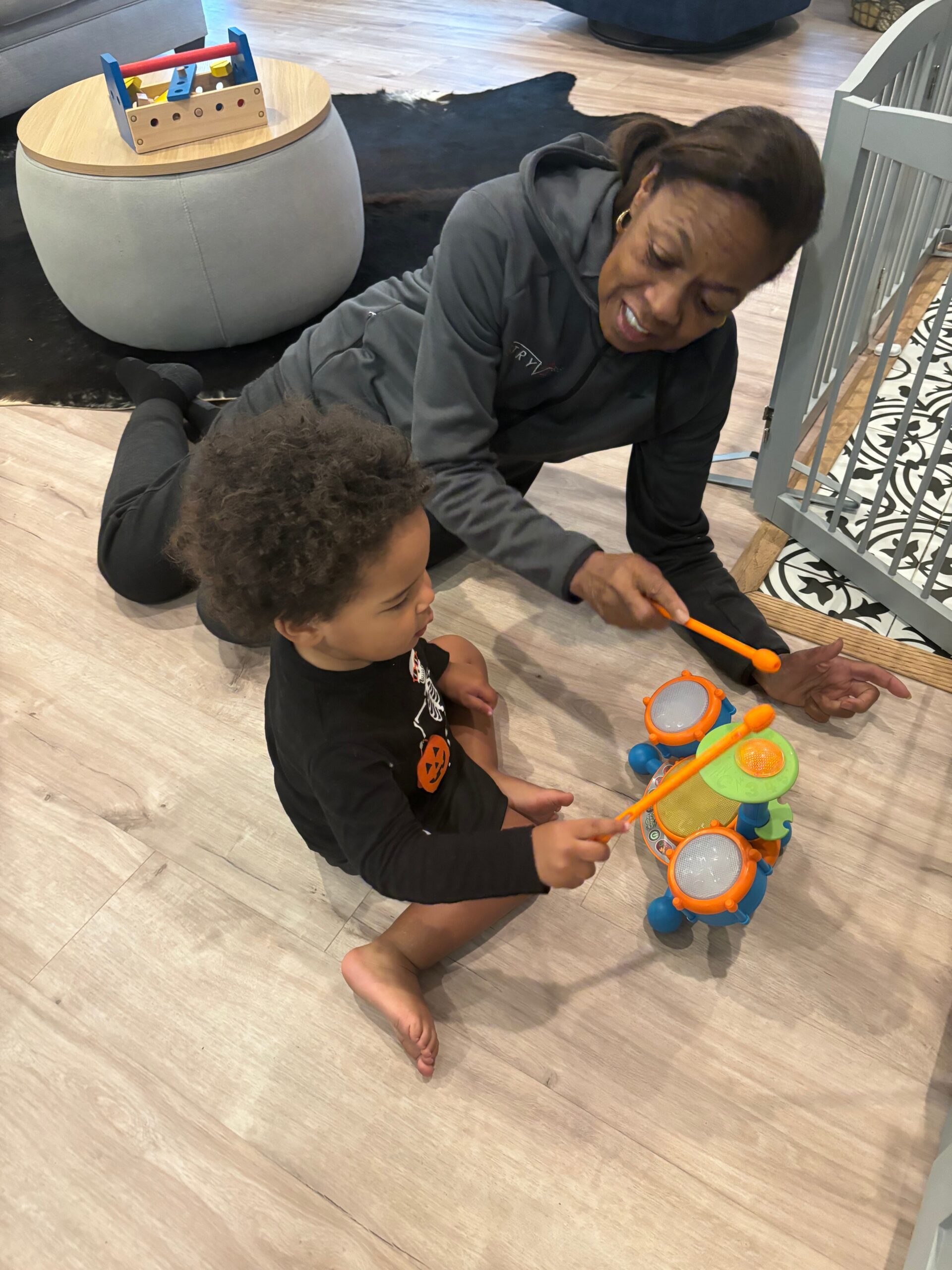 A woman and child sitting on the floor playing with a colorful toy drum set and orange drumsticks in a home setting.