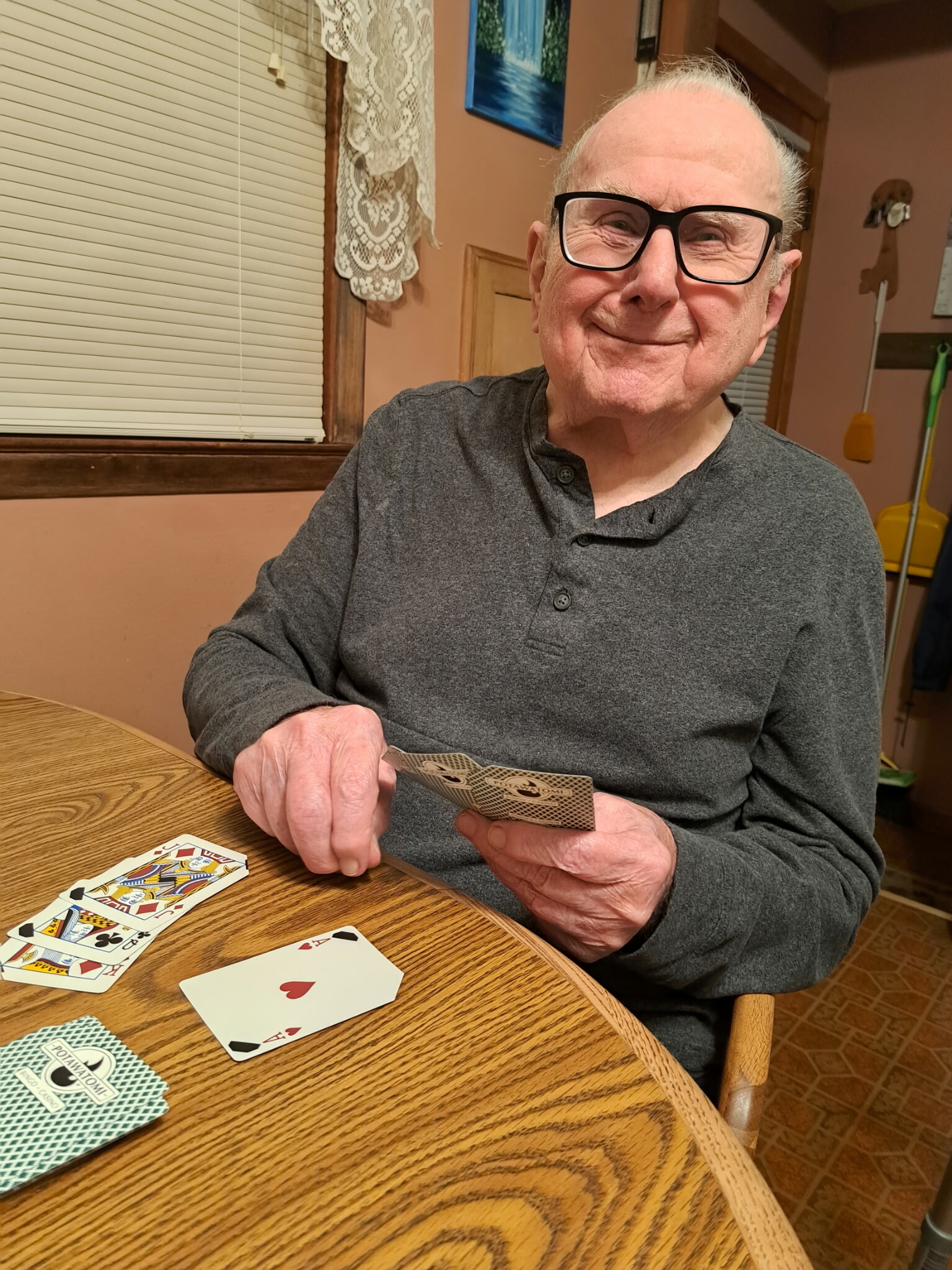 Older man with glasses sits at a wooden table, holding playing cards during a casual card game, with cards spread on the table nearby.