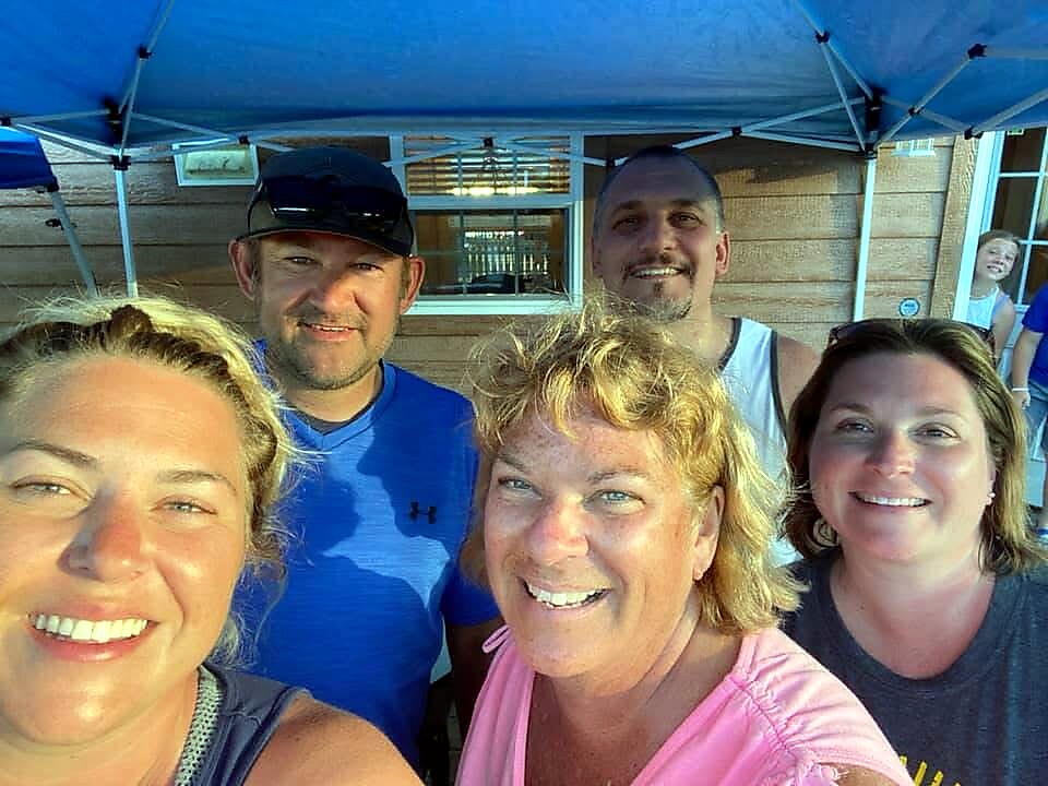 Five adults pose for a selfie under a blue canopy, smiling at the camera at an outdoor event.