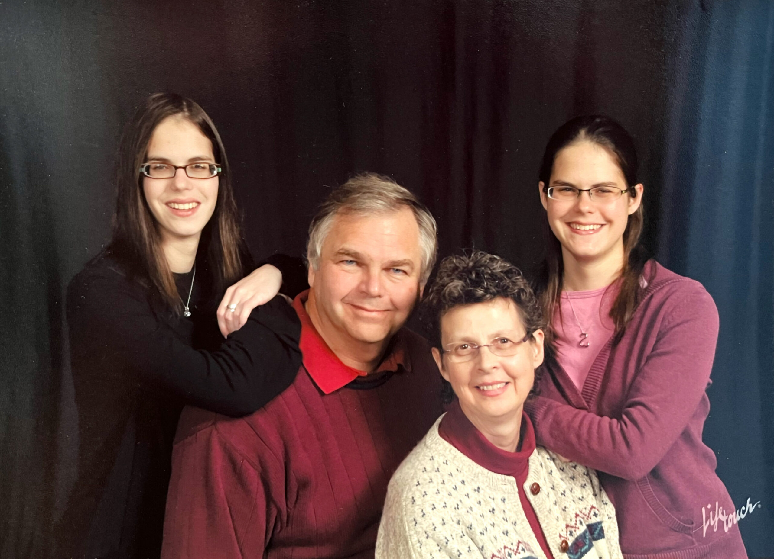 A multi-generational group of five people posing together and smiling for a portrait against a dark backdrop.