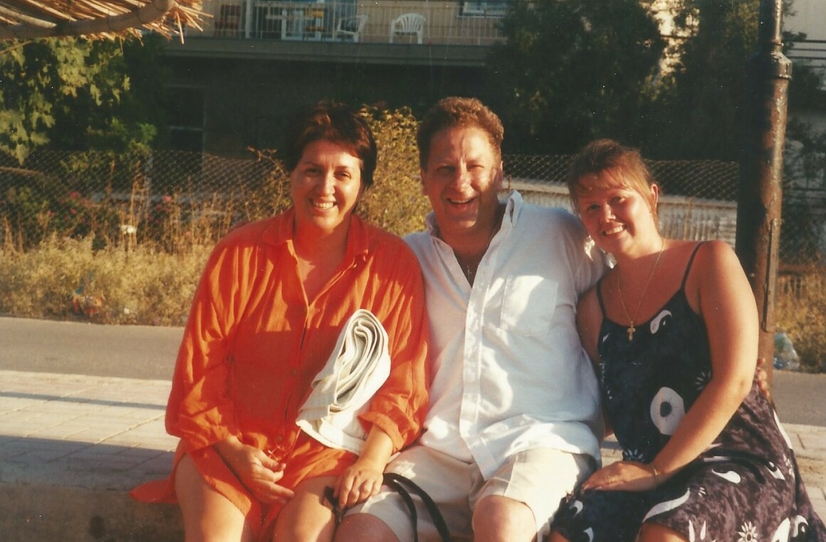 Three adults posing together on a sunny day, sitting side by side and smiling at the camera; woman in orange on the left, man in a white shirt center, woman in a black dress on the right.