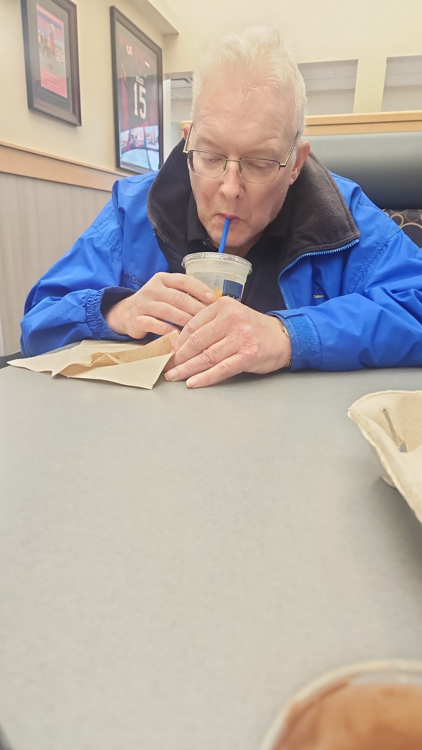 Older man wearing glasses and a blue jacket sipping a drink through a straw at a table, napkins in front, in a casual eatery.