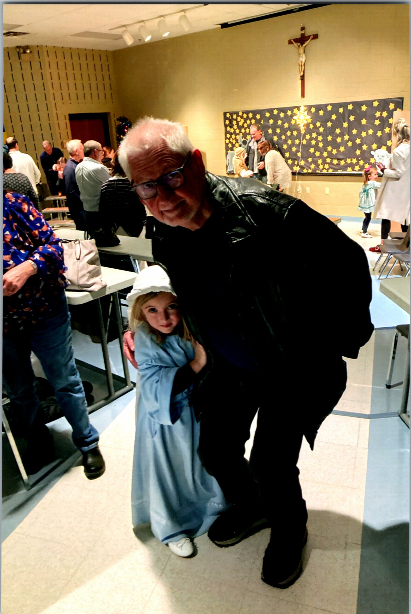 An older man in a black jacket leans toward a smiling young girl in a blue dress, indoors at a community event with a crucifix on the wall and a starry display in the background.