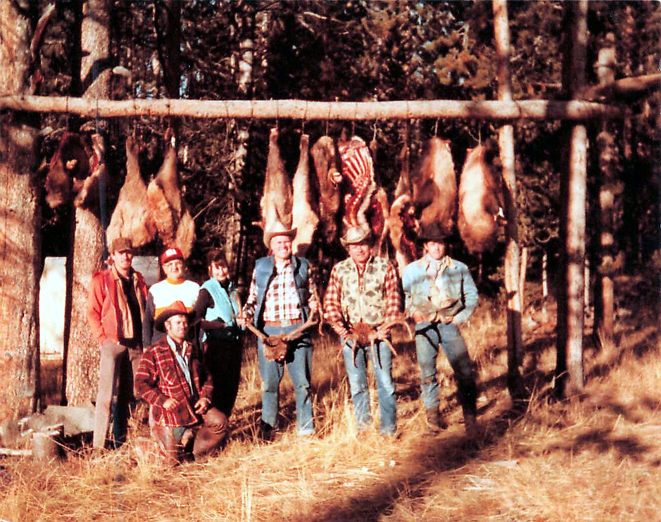 Group of people in cowboy hats and plaid shirts posing in front of a rack of animal hides outdoors in a wooded area.