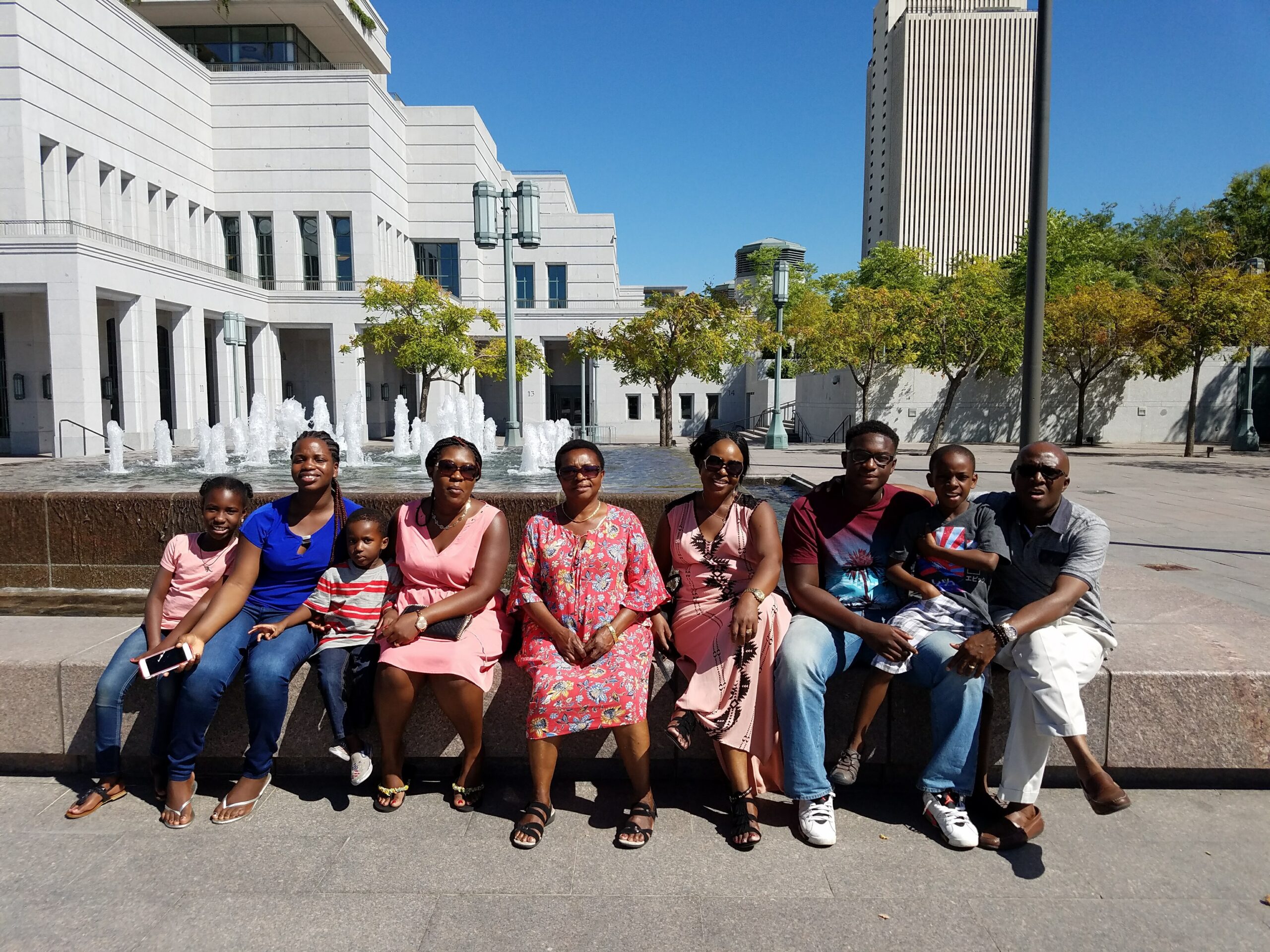 Group of people sitting along a fountain ledge outdoors in front of a modern white building on a sunny day, smiling for the photo.