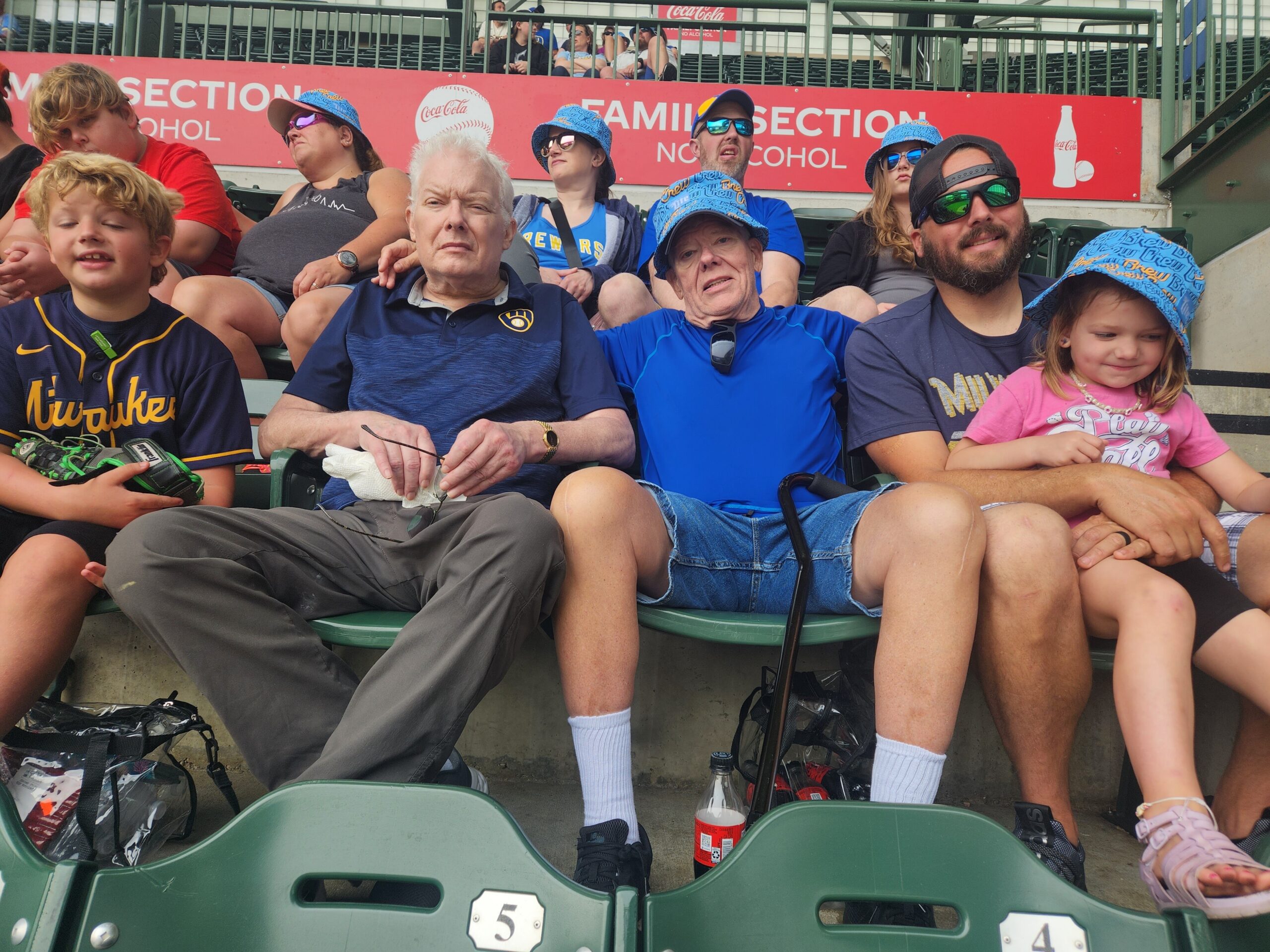 Group of diverse fans seated in stadium bleachers, wearing Brewers gear, with a Coca-Cola ad in the background near the top railing.