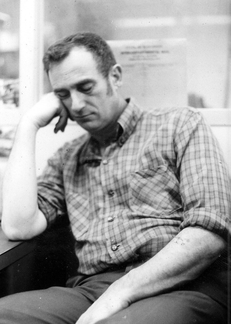Black-and-white portrait of a man wearing a plaid shirt, resting his head on his hand and looking down in thought.
