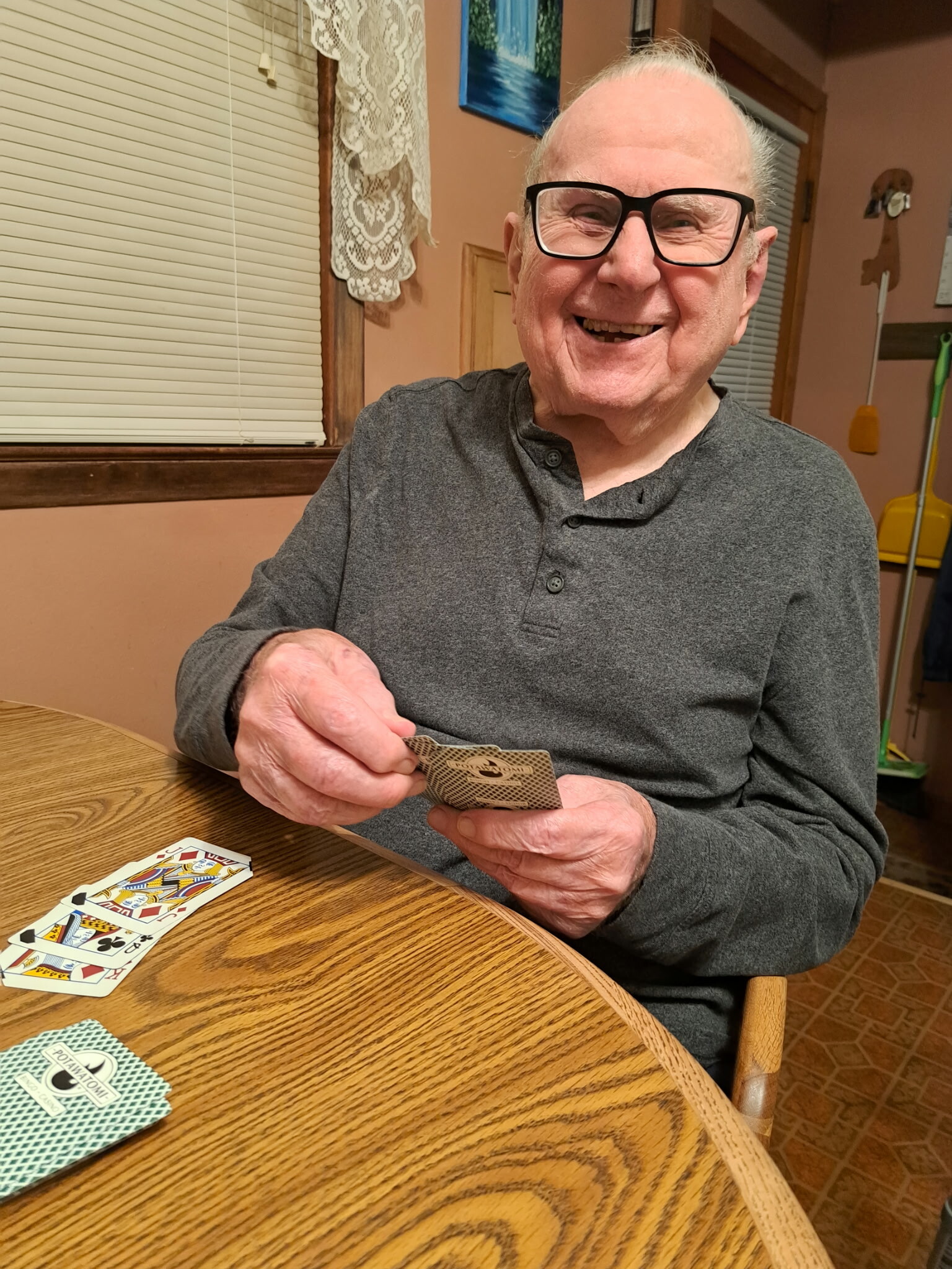 Smiling elderly man with black glasses holds playing cards at a wooden table in a cozy room.