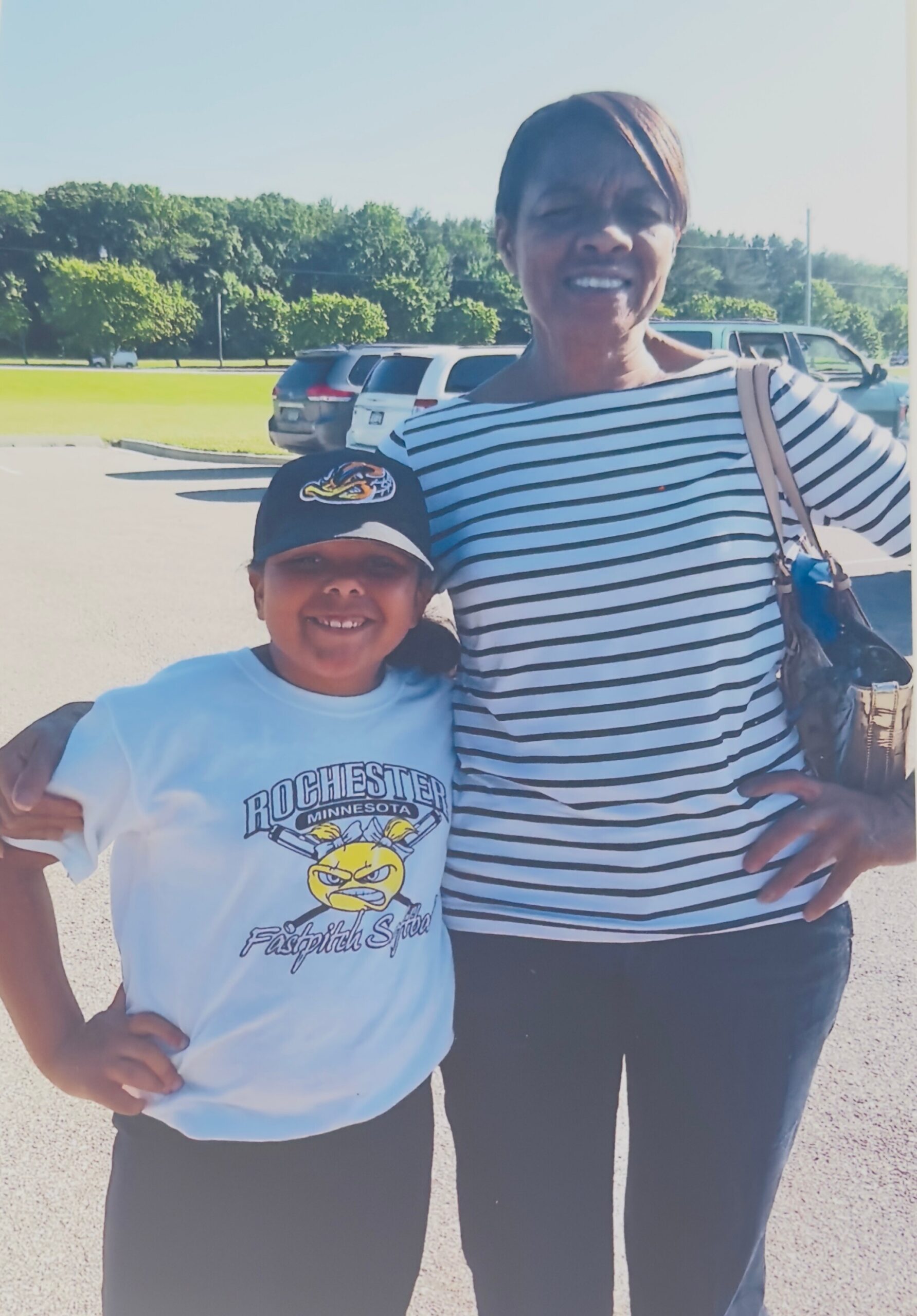 Two people pose for a sunny outdoor photo in a parking lot: a woman in a striped top and a girl in a Rochester, Minnesota softball shirt with a cap, both smiling and arms around each other.