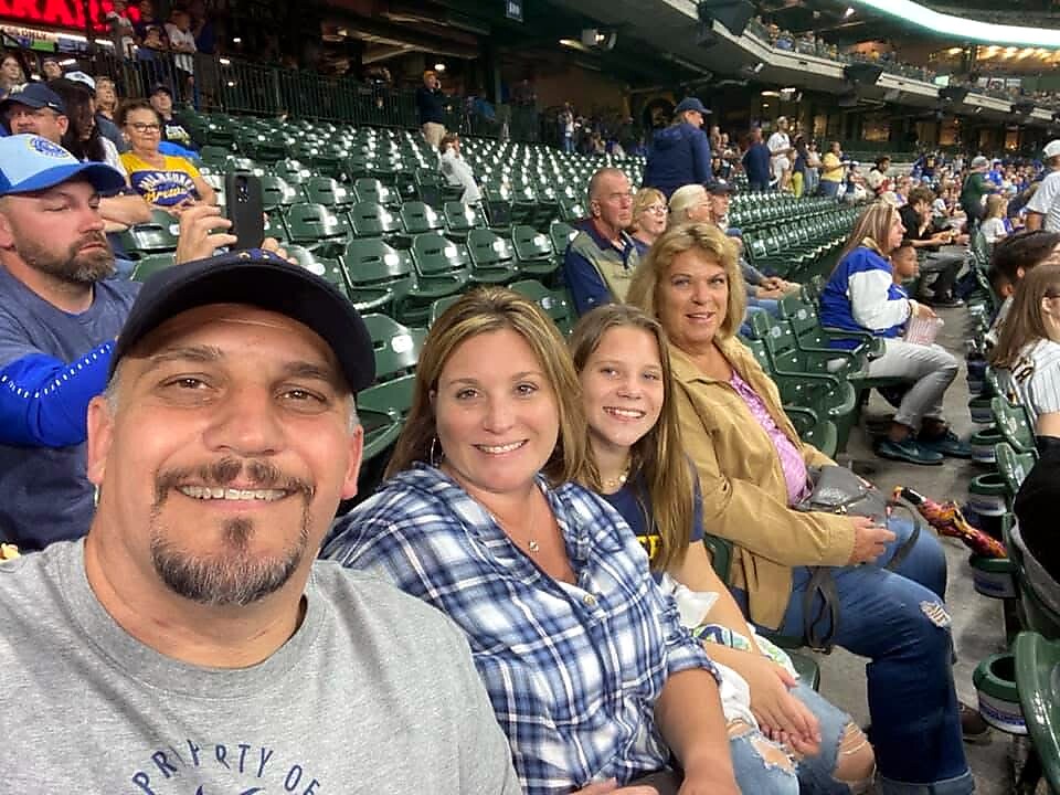 Man in a baseball cap taking a selfie with two women and a girl in stadium seating at a game; smiles at the camera with fans in the background.