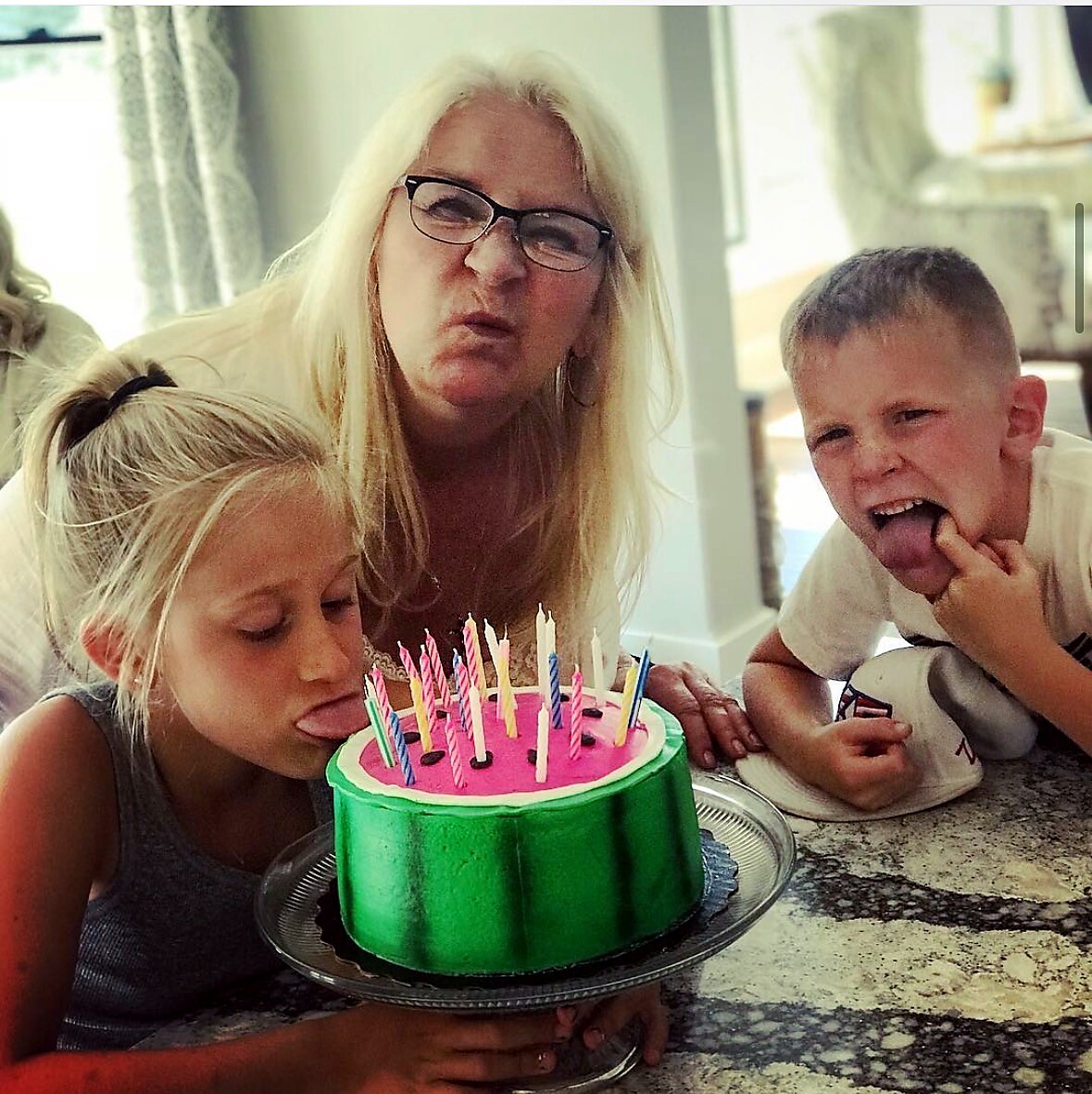 A woman with two children before a pink-trosted cake with candles, the girl licking the cake while the boy makes a goofy face nearby.