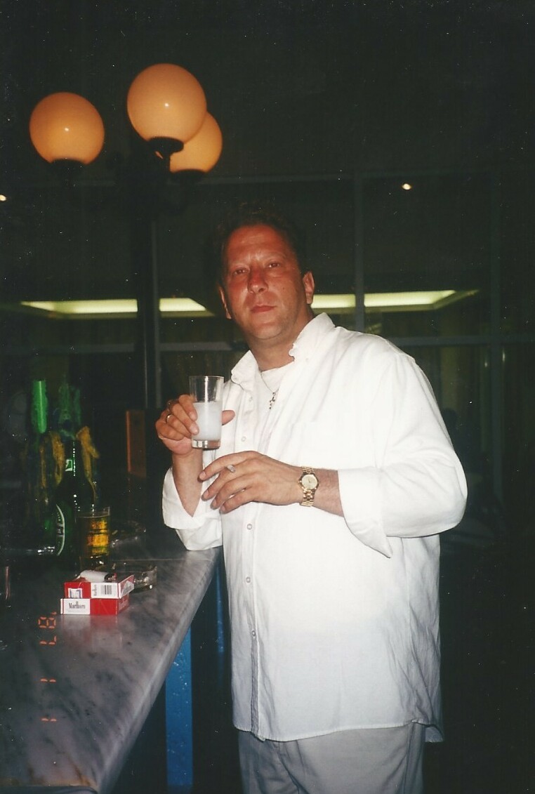 Man in a white shirt stands at a bar, holding a glass in a toast with a watch on his wrist, bottles visible in the background.