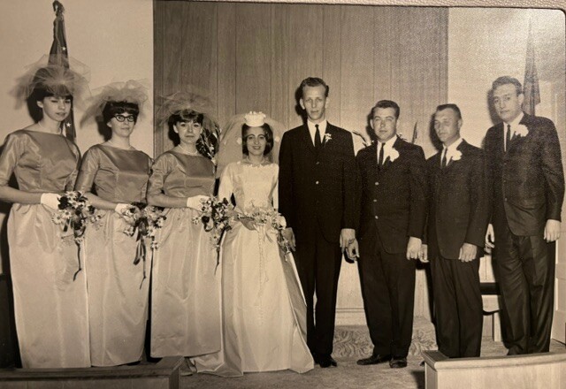 A bride in a white gown stands with bridesmaids on the left and groomsmen on the right during a wedding ceremony photo in a theater setting.