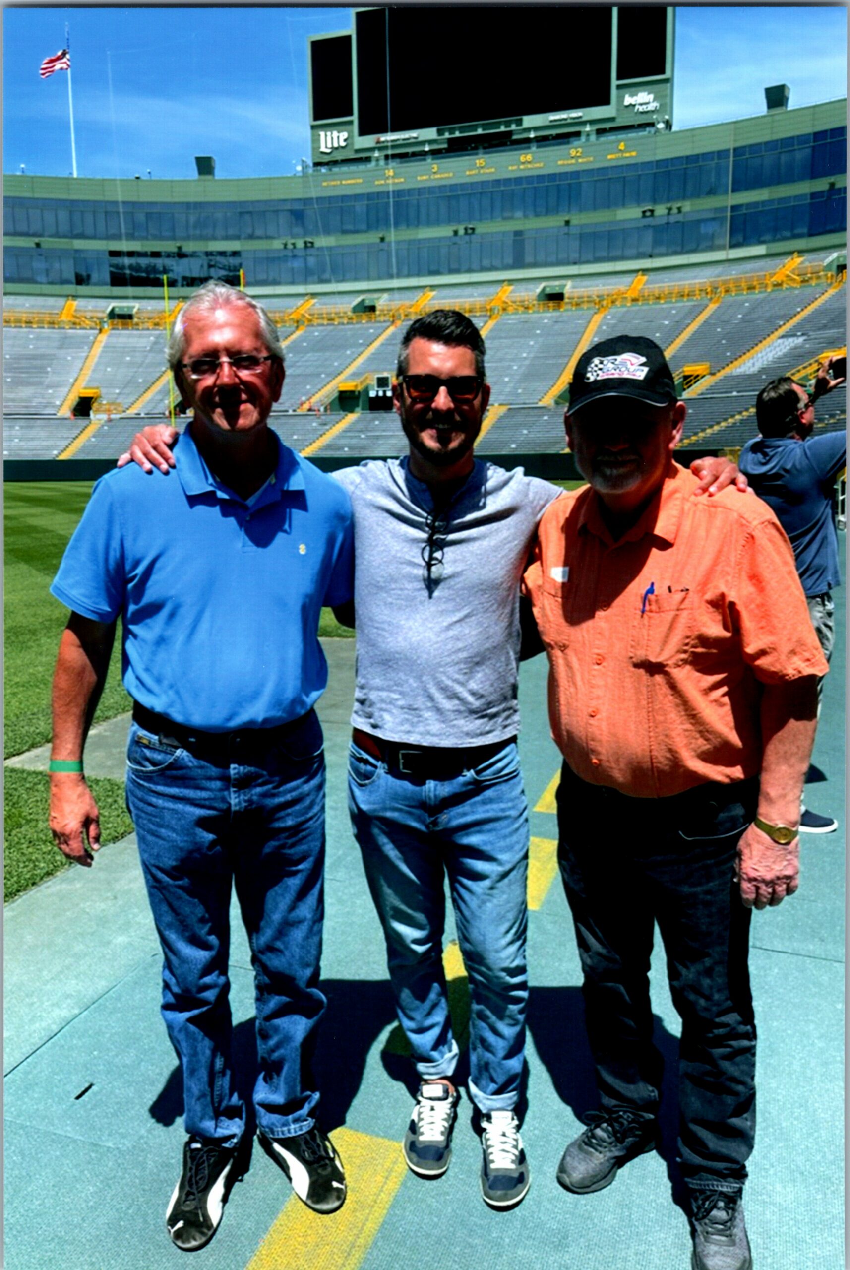 Three men stand together with arms around each other on a football field, stadium seating and a large scoreboard behind them, American flag in the breeze.