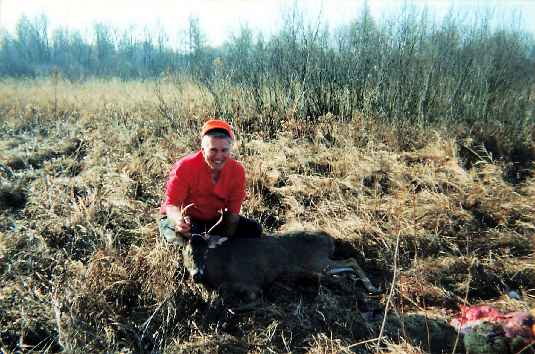 Man in red jacket and orange cap kneels in a field of tall dry grass, posing with a dark animal carcass at his feet during a hunt.