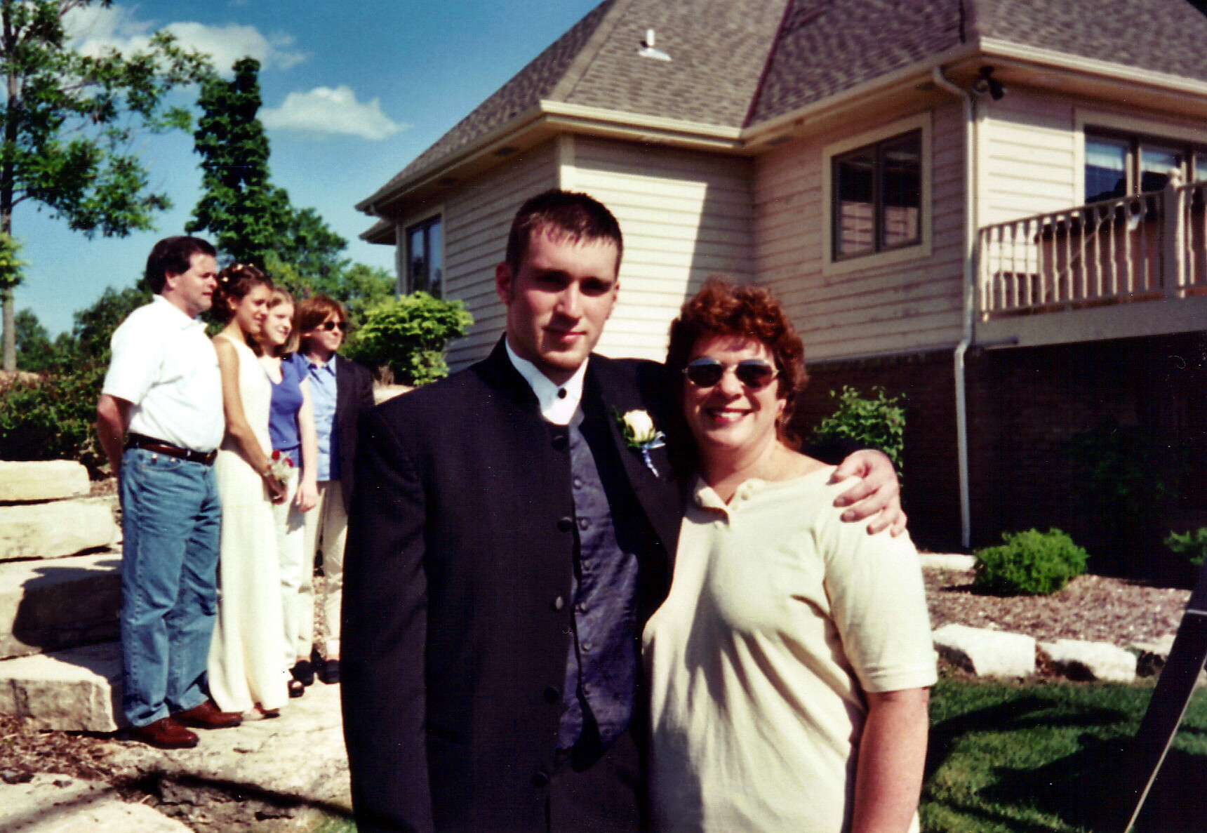 Young man in a dark suit with a boutonniere stands beside a smiling woman in a pale yellow top, outdoors in front of a house.