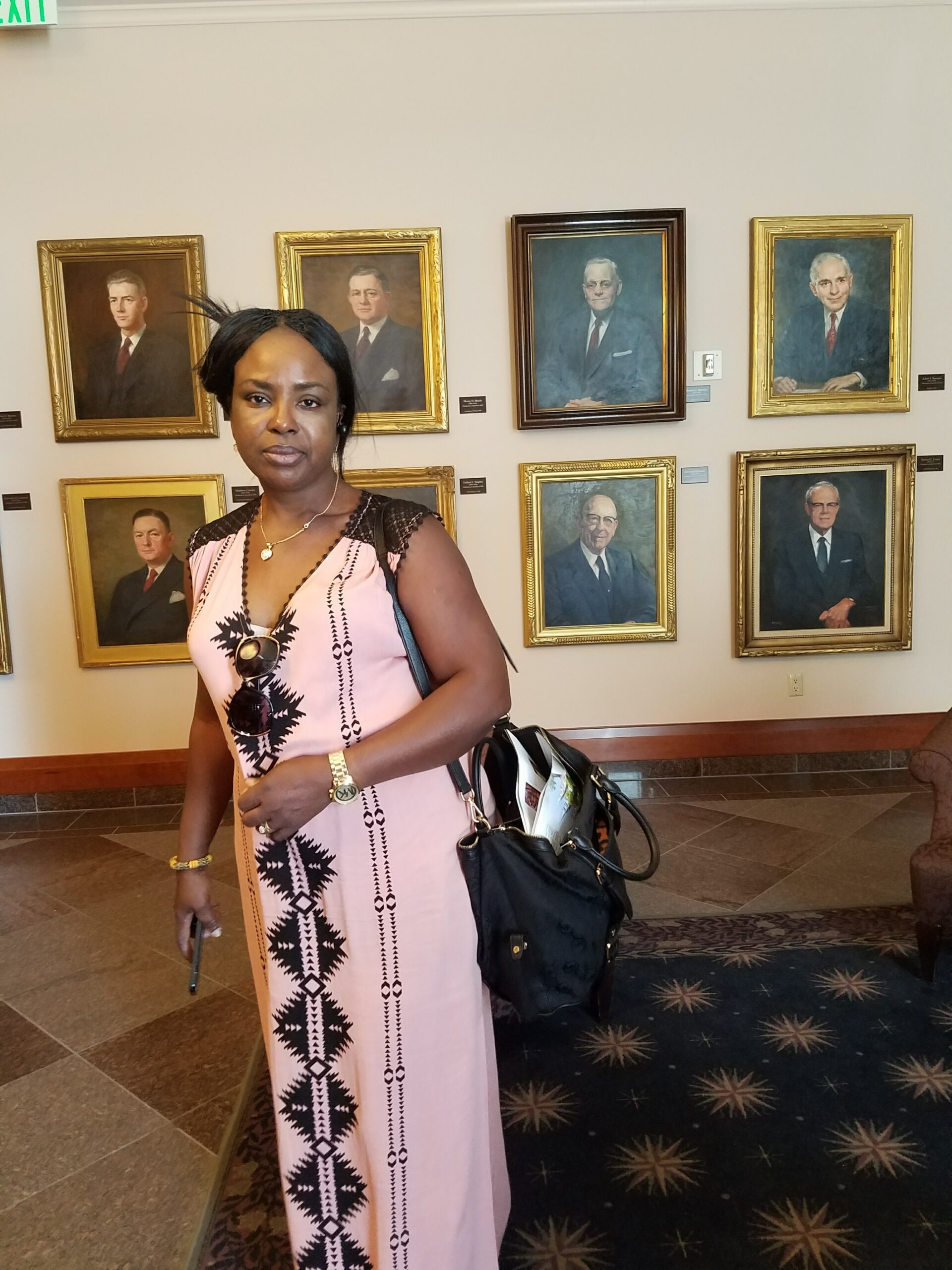 Woman in a pink patterned dress stands in a gallery, in front of framed portraits on a beige wall, with a black bag and sunglasses hanging from her dress