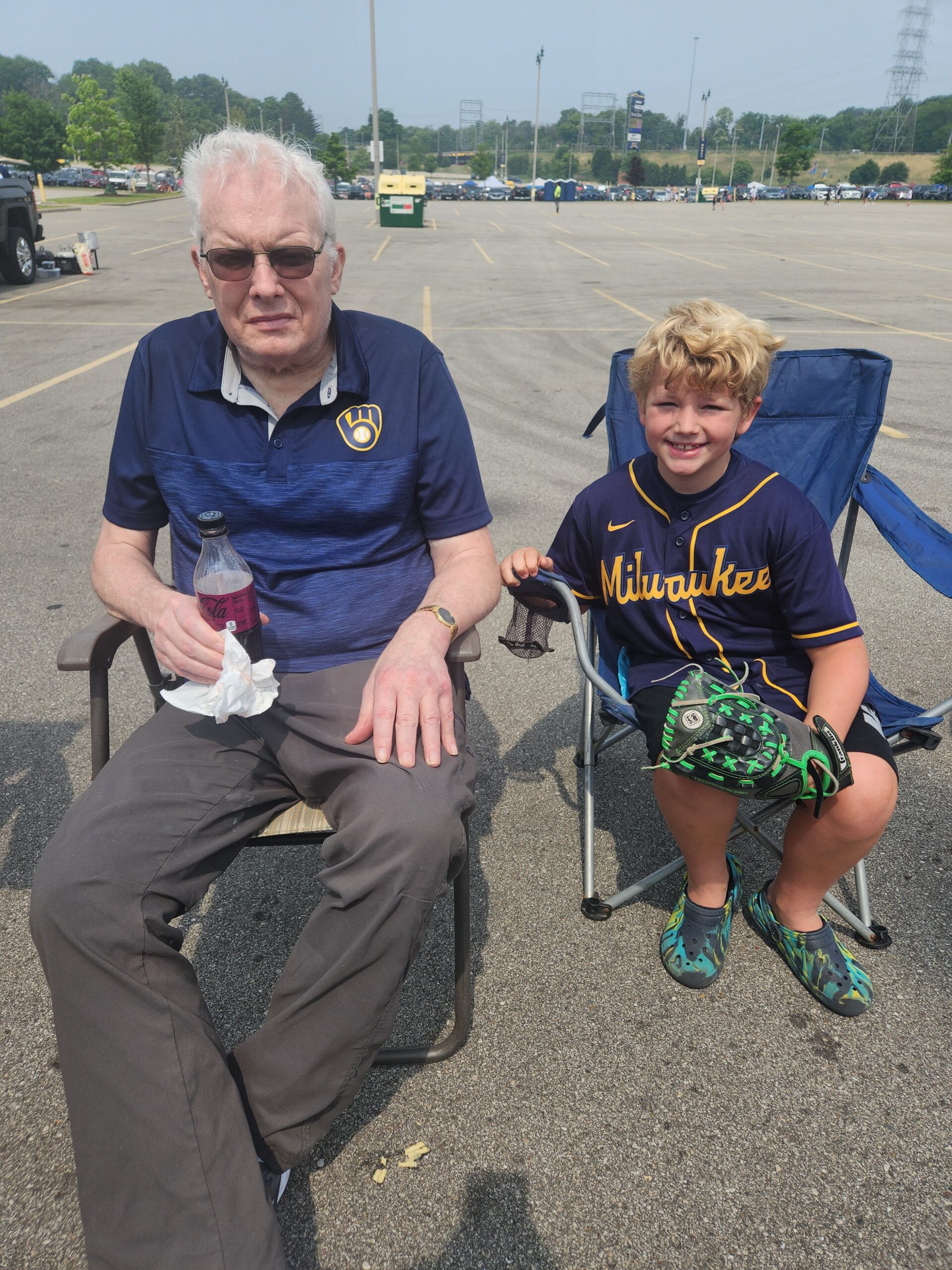 Older man with white hair and sunglasses sits in a folding chair in a parking lot, holding a pink soda bottle; a smiling boy in a Milwaukee Brewers jersey sits beside him in a blue chair with a baseball glove nearby.
