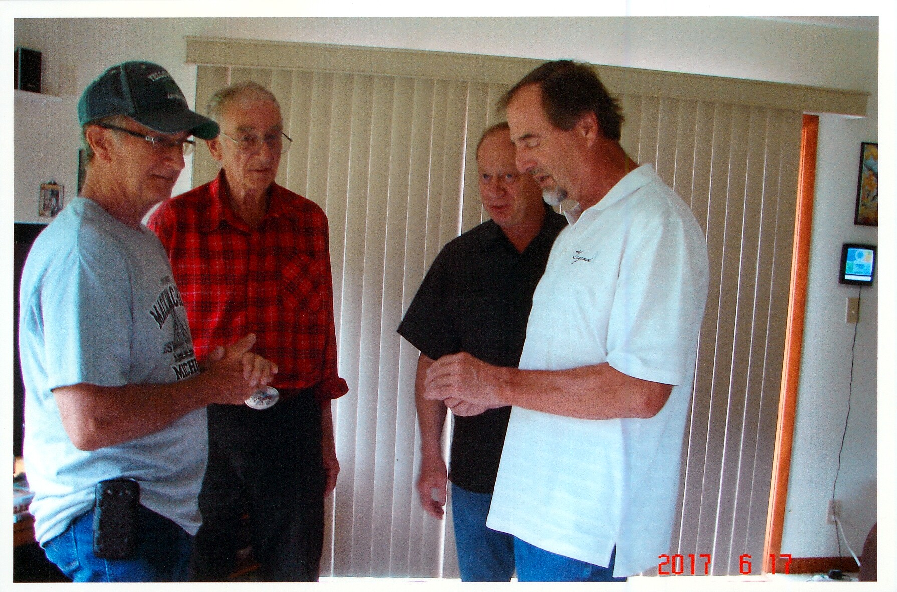Four men in casual clothing stand together in a living room by vertical blinds, examining a small round object held by the man in the light blue shirt.