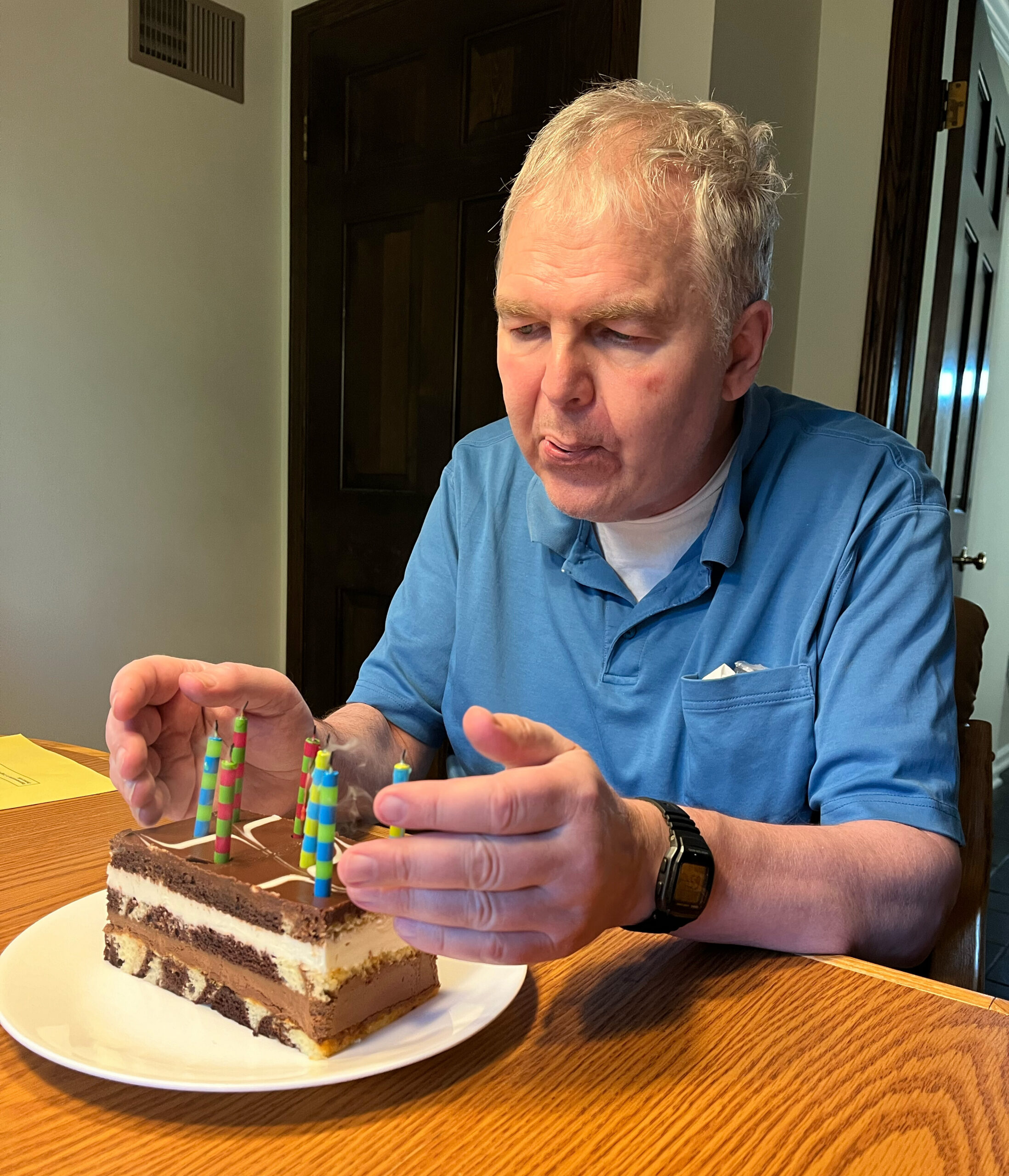 Man in a blue shirt sits at a wooden table, hands near a layered chocolate cake with rainbow-striped candles, indoors.
