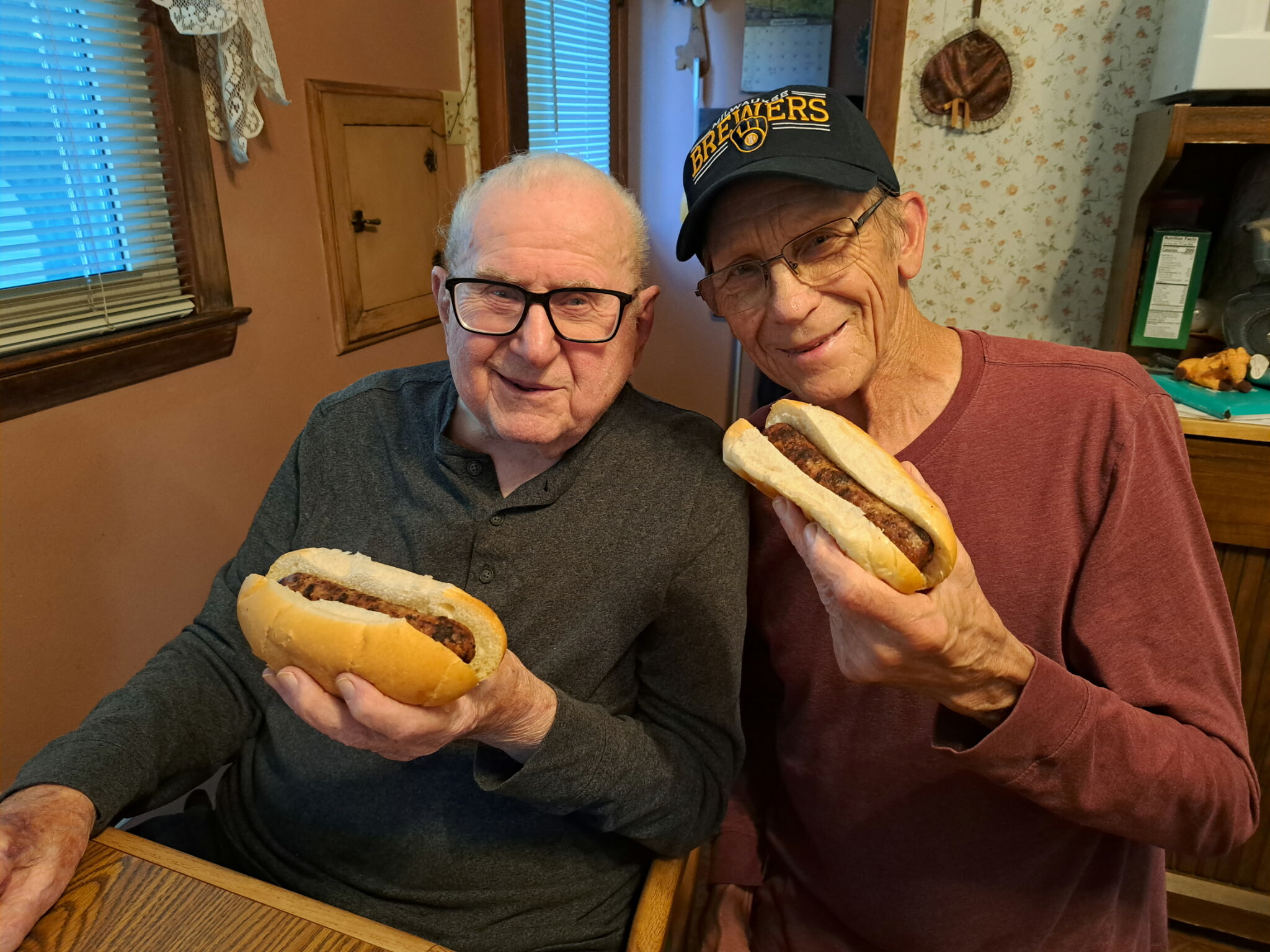 Two elderly men with glasses smiling, each holding a hot dog in a kitchen.