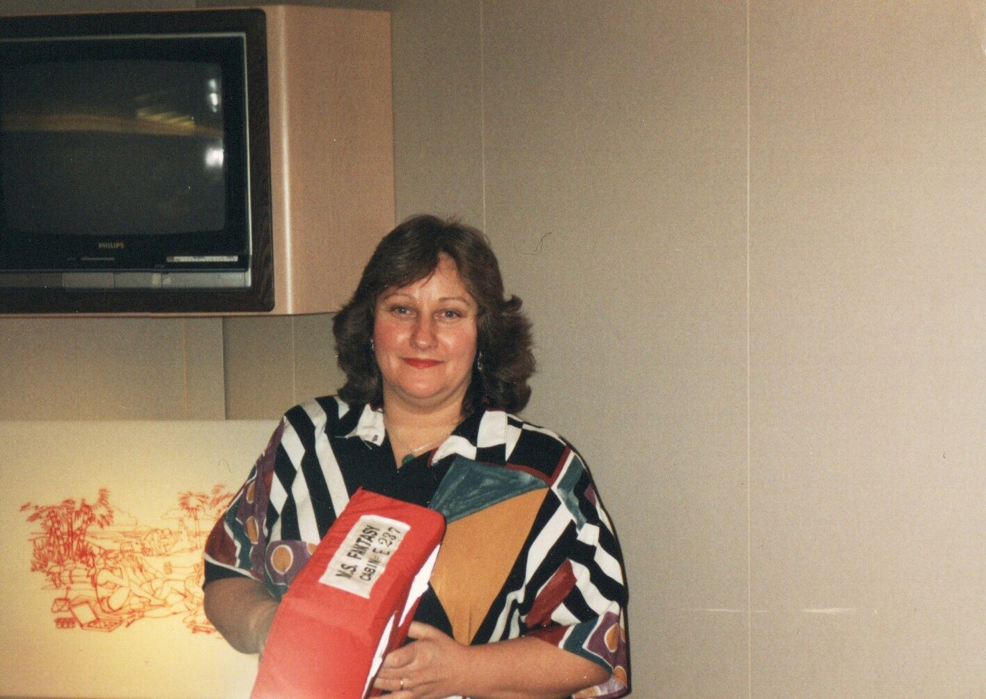 Smiling woman with shoulder-length hair in a colorful geometric blouse, holding a red folder or portfolio in a kitchen-like room with a retro TV above.