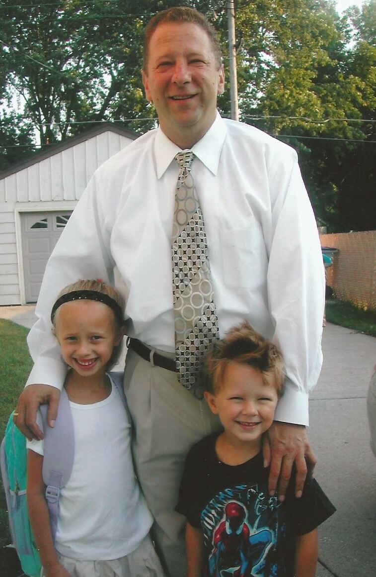 Man in a white shirt and patterned tie stands with two smiling children outside a house.