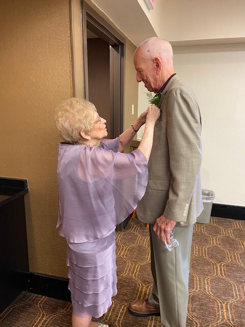 Elderly woman pins a boutonniere on an elderly man's gray suit in a hallway.