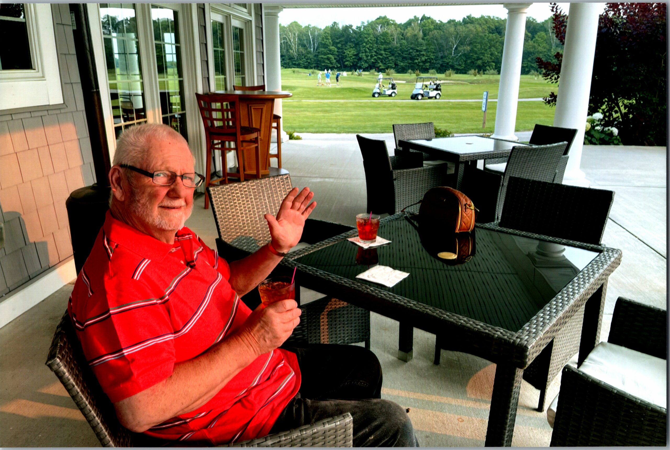 Older man in a red striped polo sits at an outdoor patio table with two cocktails, waves to the camera as a golf course sits beyond.