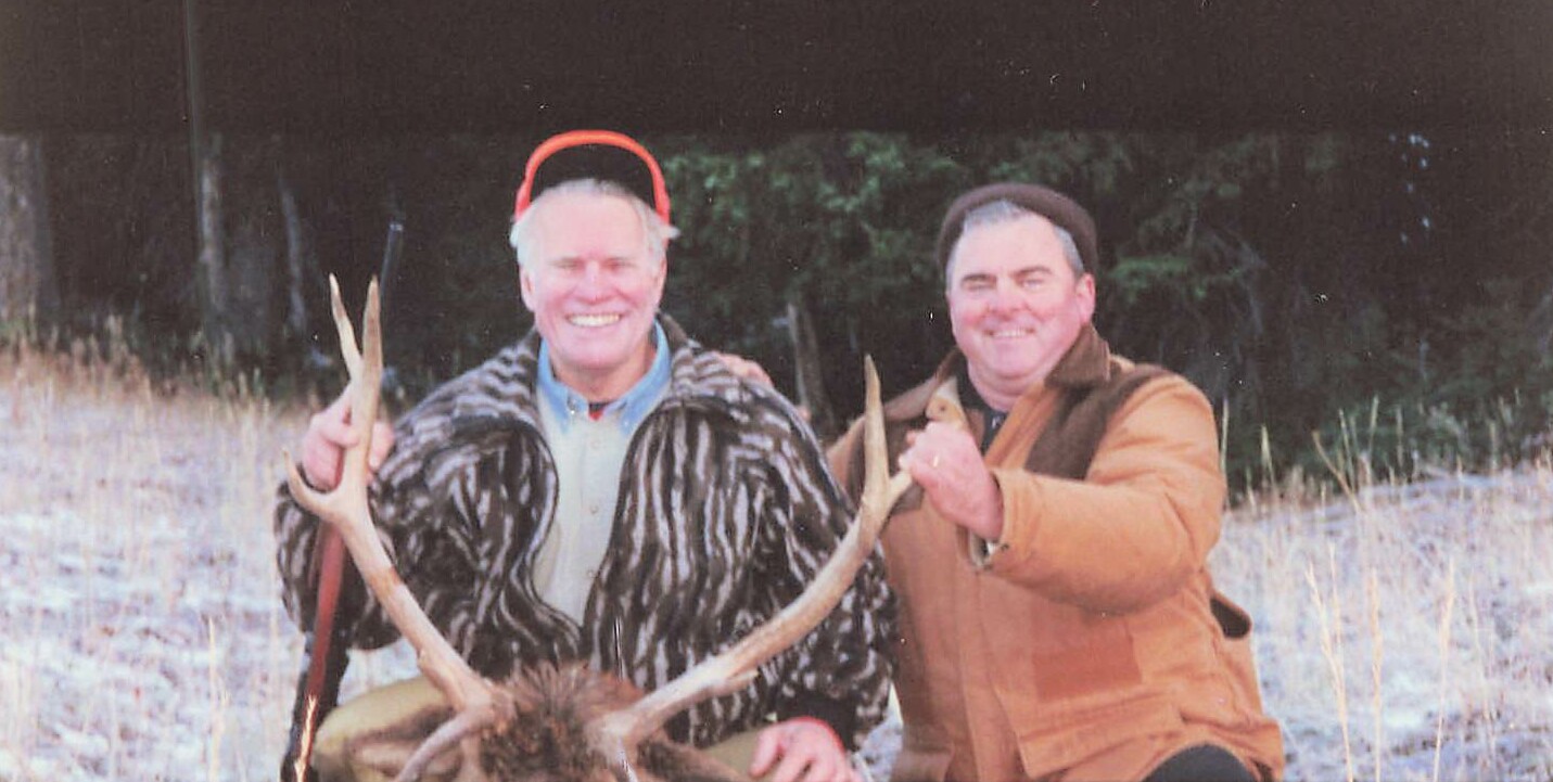 Two men posing outdoors with a large elk antlered head, one wearing a fur coat and red hat, the other in a brown jacket, in a grassy field at edge of trees.