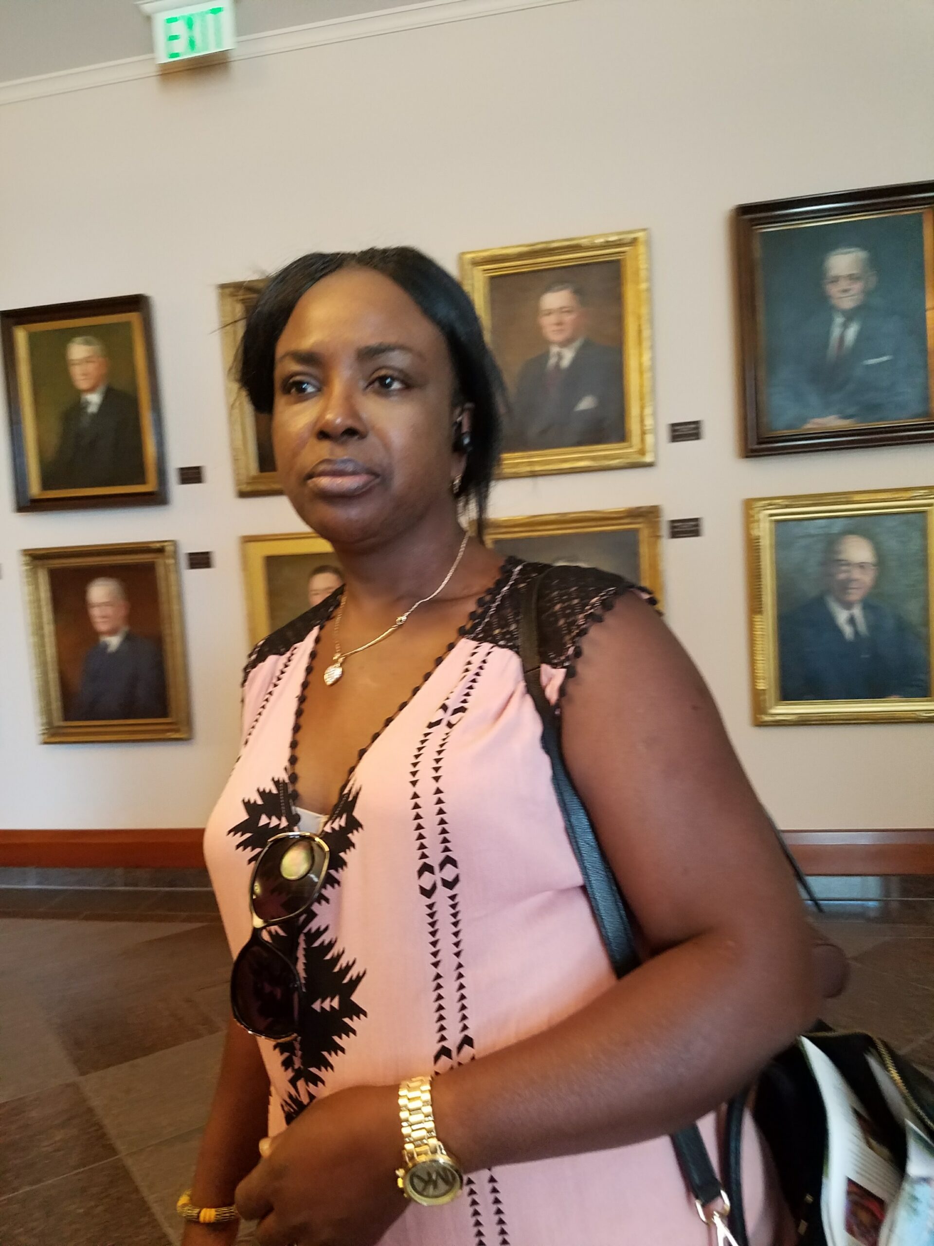 African American woman in a pink patterned top stands in an art gallery with gold-framed portraits on the wall behind her.
