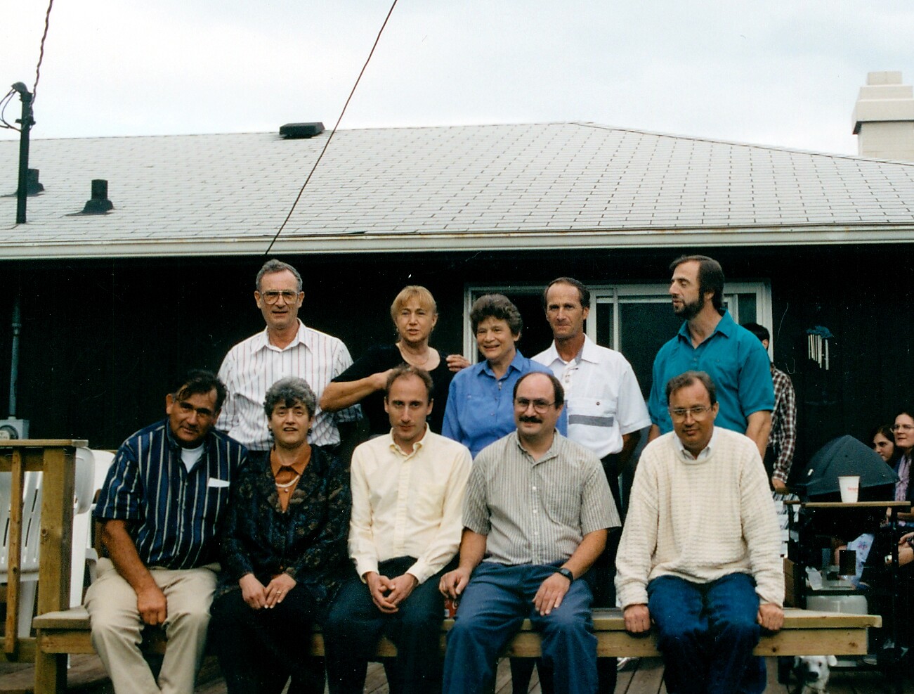 A large group of adults posing for a casual photo on a wooden bench and deck in a backyard, with a house and sloped roof in the background.