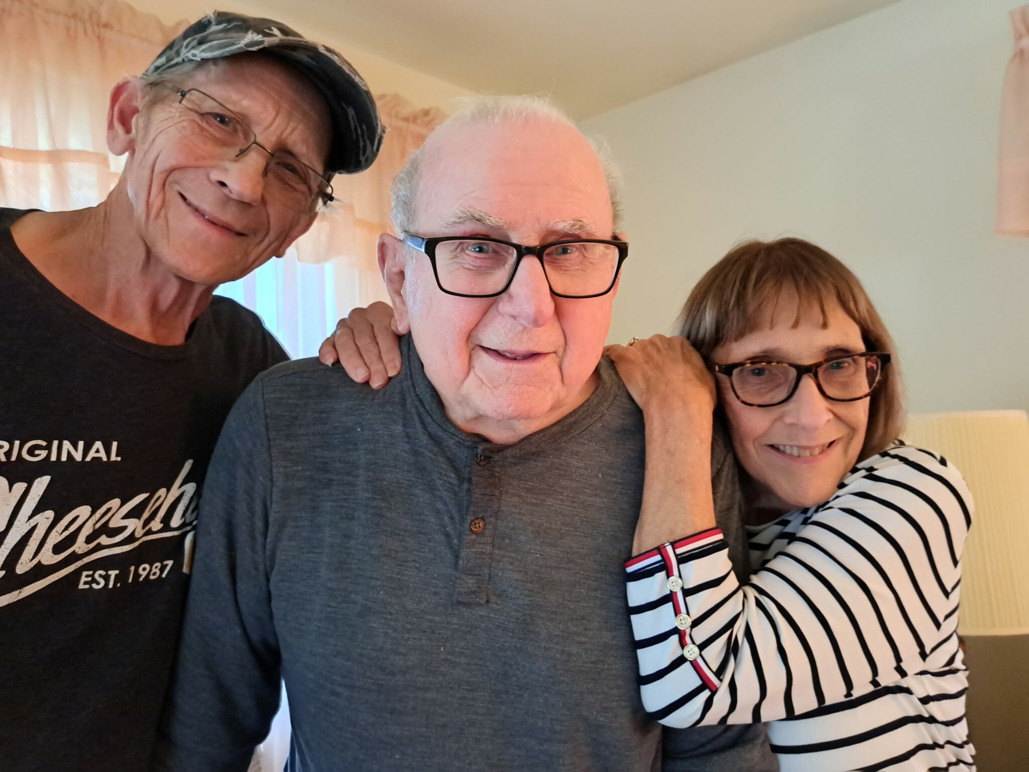 Three adults hug and smile at the camera in a cozy indoor setting, arms around each other.