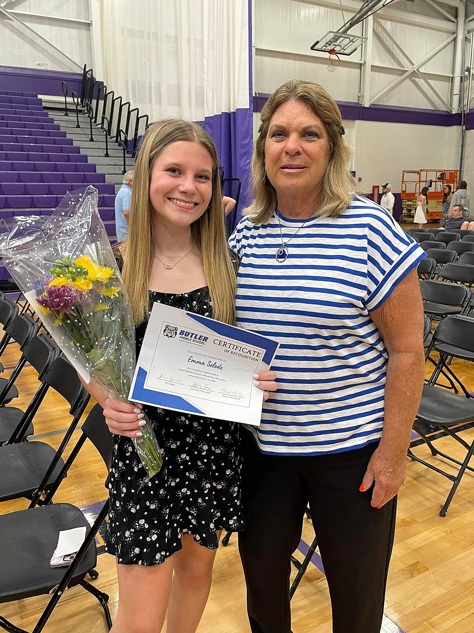 Smiling teenage girl in a black floral dress holds a bouquet of flowers and a certificate, standing with an older woman in a blue-and-white striped shirt at a school gymnasium.