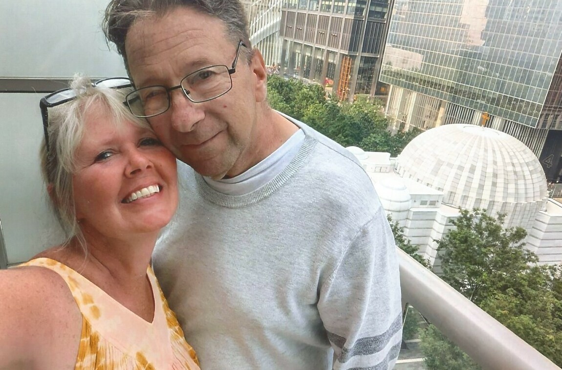 Smiling couple posing for a selfie on a balcony with city skyscrapers and a domed building in the background.