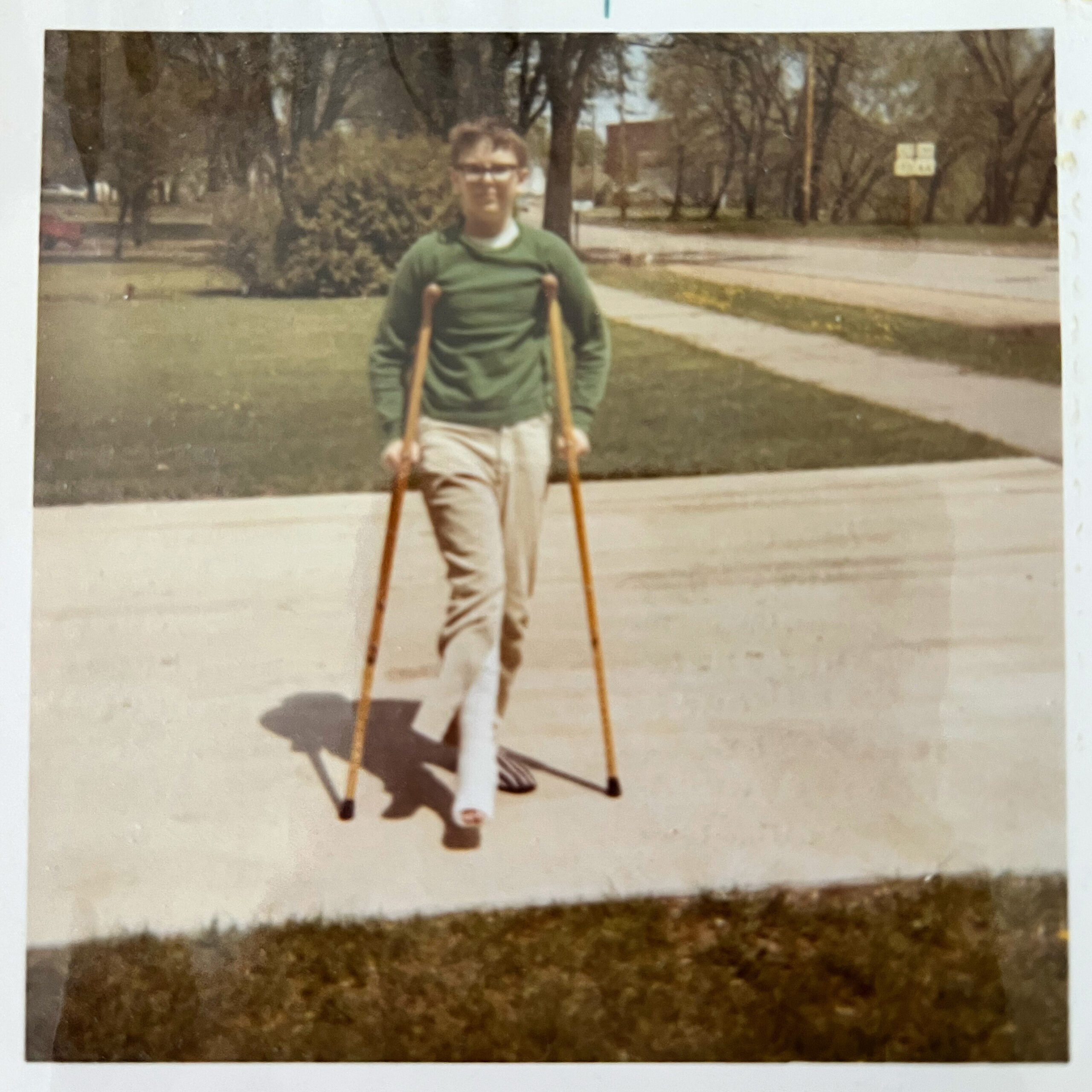 Man using crutches outside in a park, wearing a green sweater and light pants.