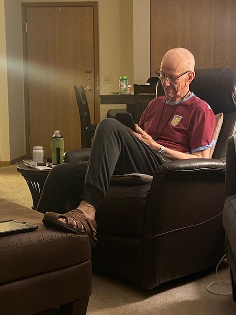 Older man with glasses sits in a leather chair, reading a tablet in a living room setting.