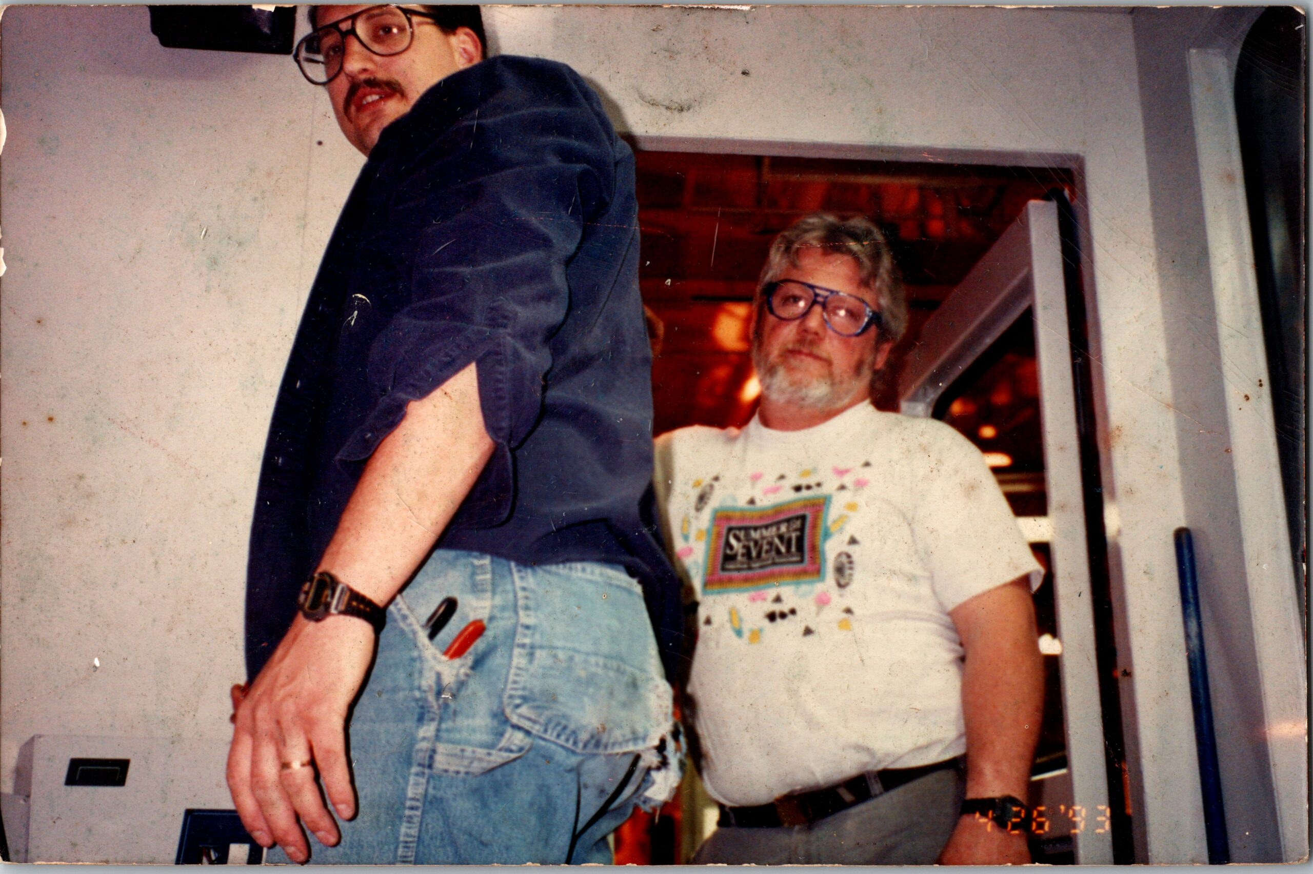 Two men pose in a doorway; foreground man in a navy shirt and jeans, with a watch, the other man wearing glasses and a graphic tee behind him.