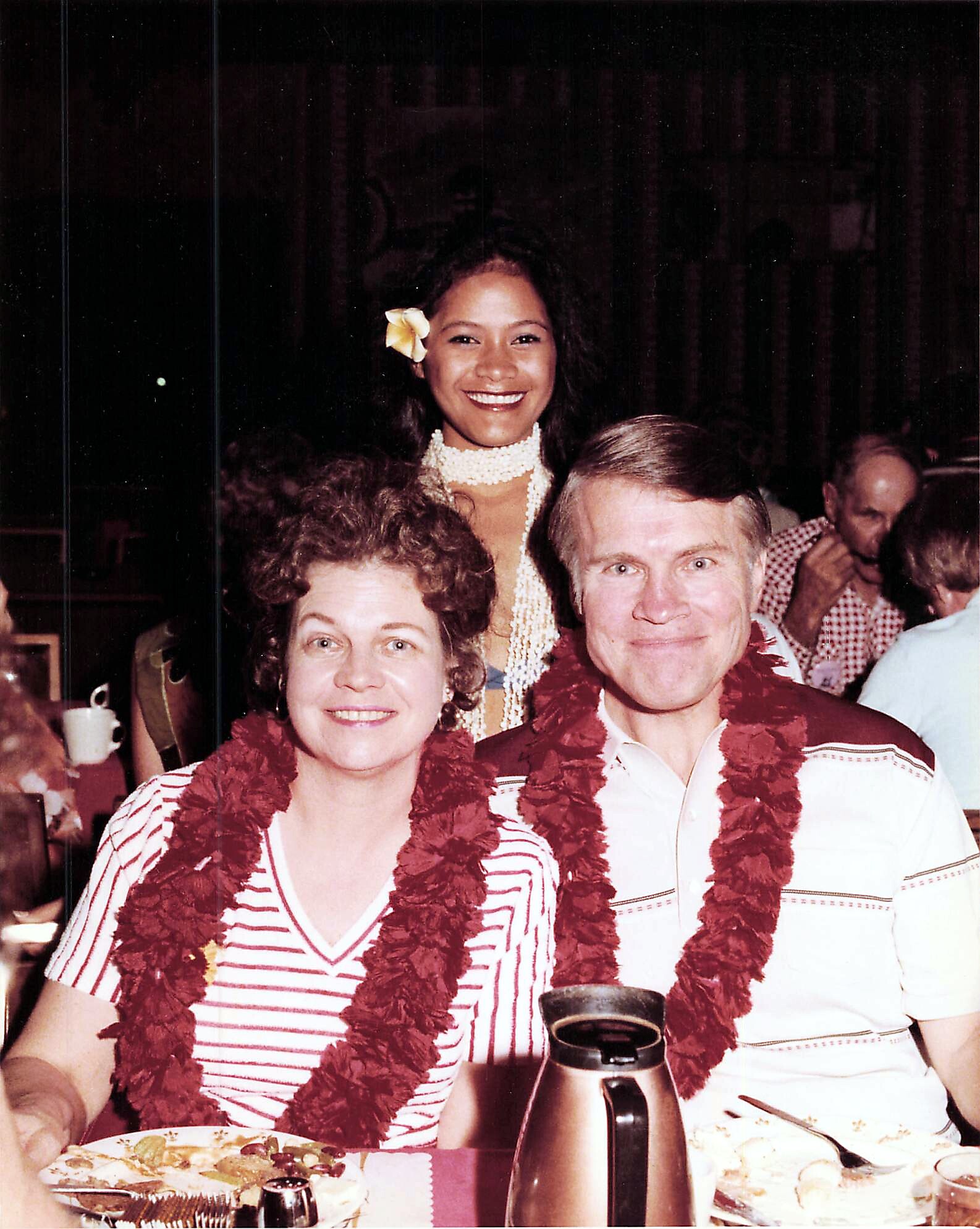 Three friends at a Hawaiian-themed party wearing leis and a flower in the hair, smiling at the camera.