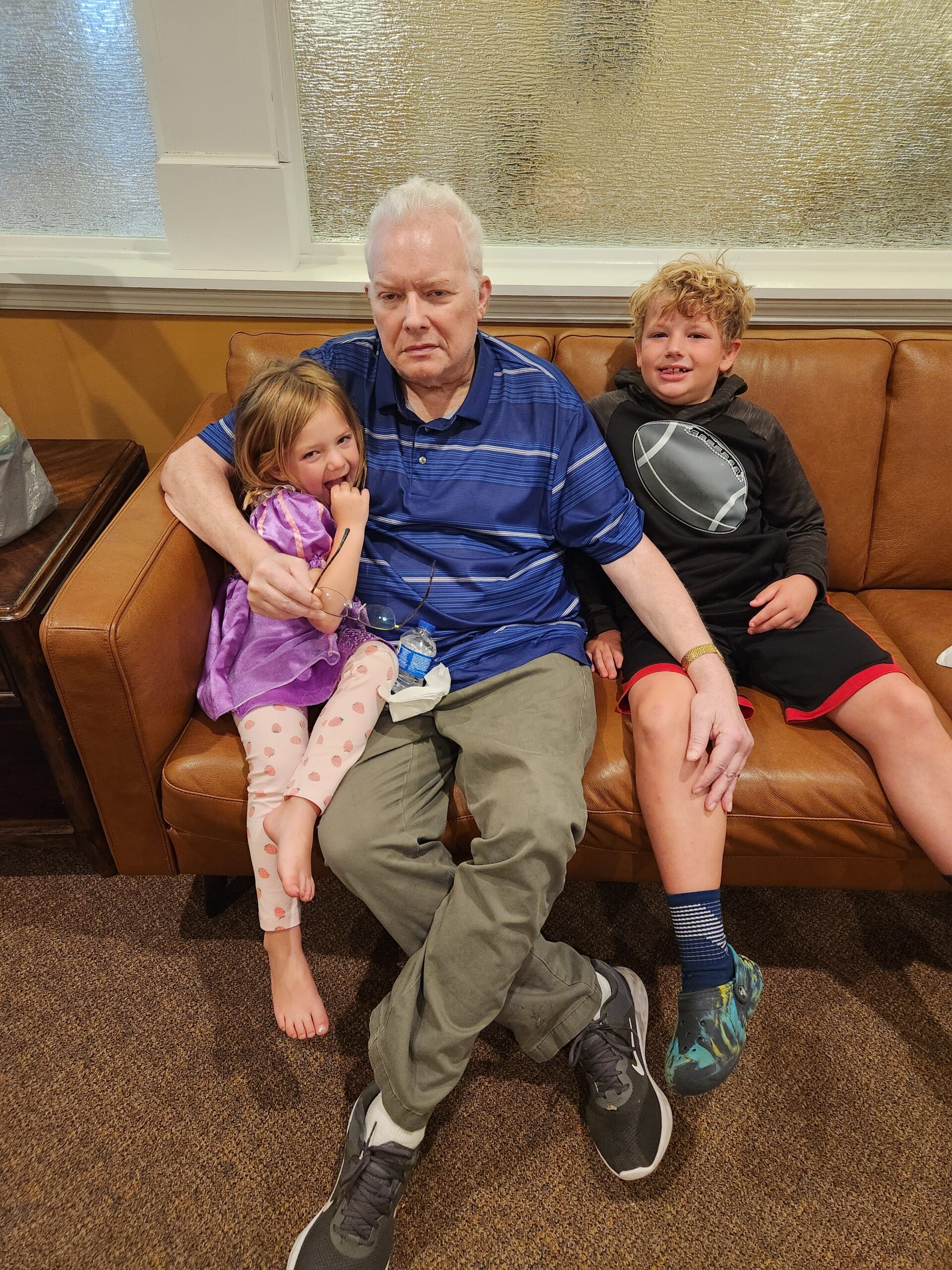 An older man sits on a brown sofa with a young girl in a purple dress snuggled beside him and a boy in a black hoodie on the other side, in a waiting room setting.