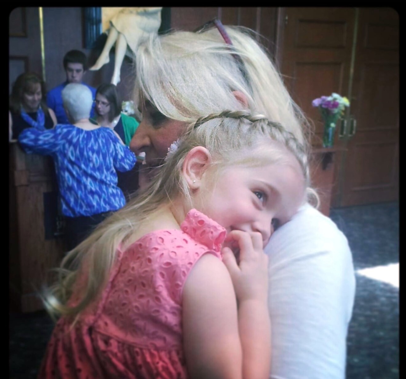Two girls share a hug in a lobby: a young girl in a pink dress hugs an older woman with braided hair, smiling in the background.