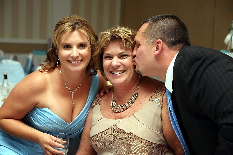 Three adults at a formal event: a woman in a blue gown on the left, a beige-dressed woman in the middle, and a man in a suit kissing her cheek on the right, all smiling.