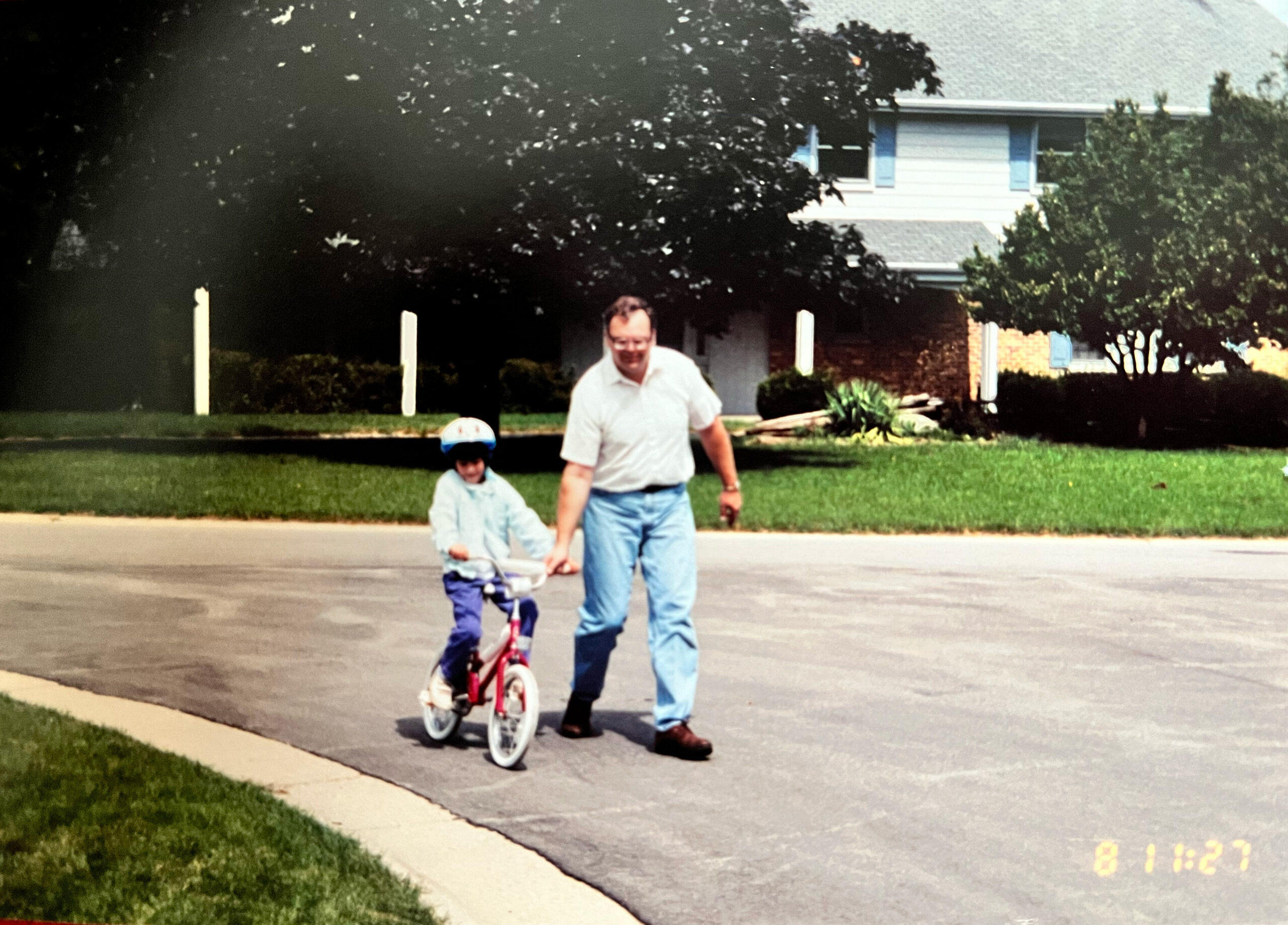 Man guiding a young child on a small bicycle along a suburban street, holding hands and smiling.